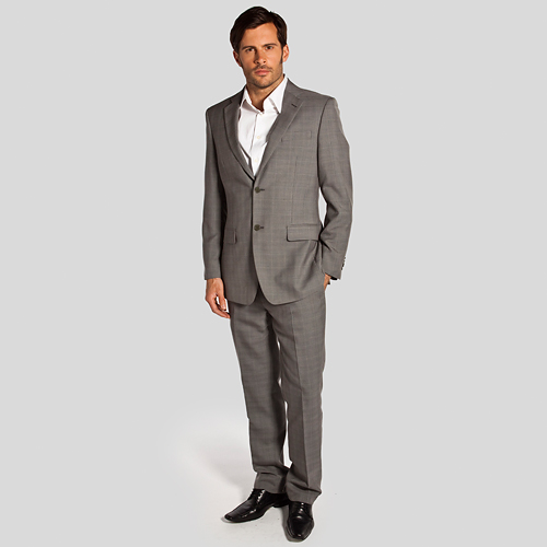 Mężczyzna w szarym garniturze w kratę, pozujący na jasnoszarym tle w Gdyni. Garnitur z dwoma guzikami, biała koszula, czarne lakierowane buty.