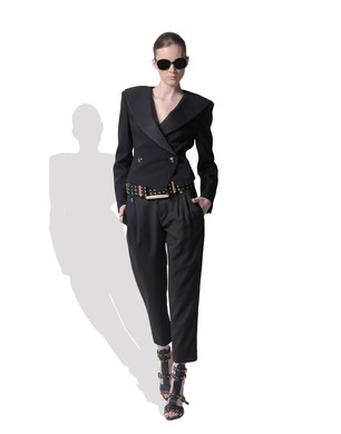 Kobieta prezentuje czarny, dwurzędowy żakiet z szerokimi ramionami, spodnie typu marchewki z paskiem nabijanym ćwiekami oraz sandały na obcasie, całość na białym tle z cieniem.