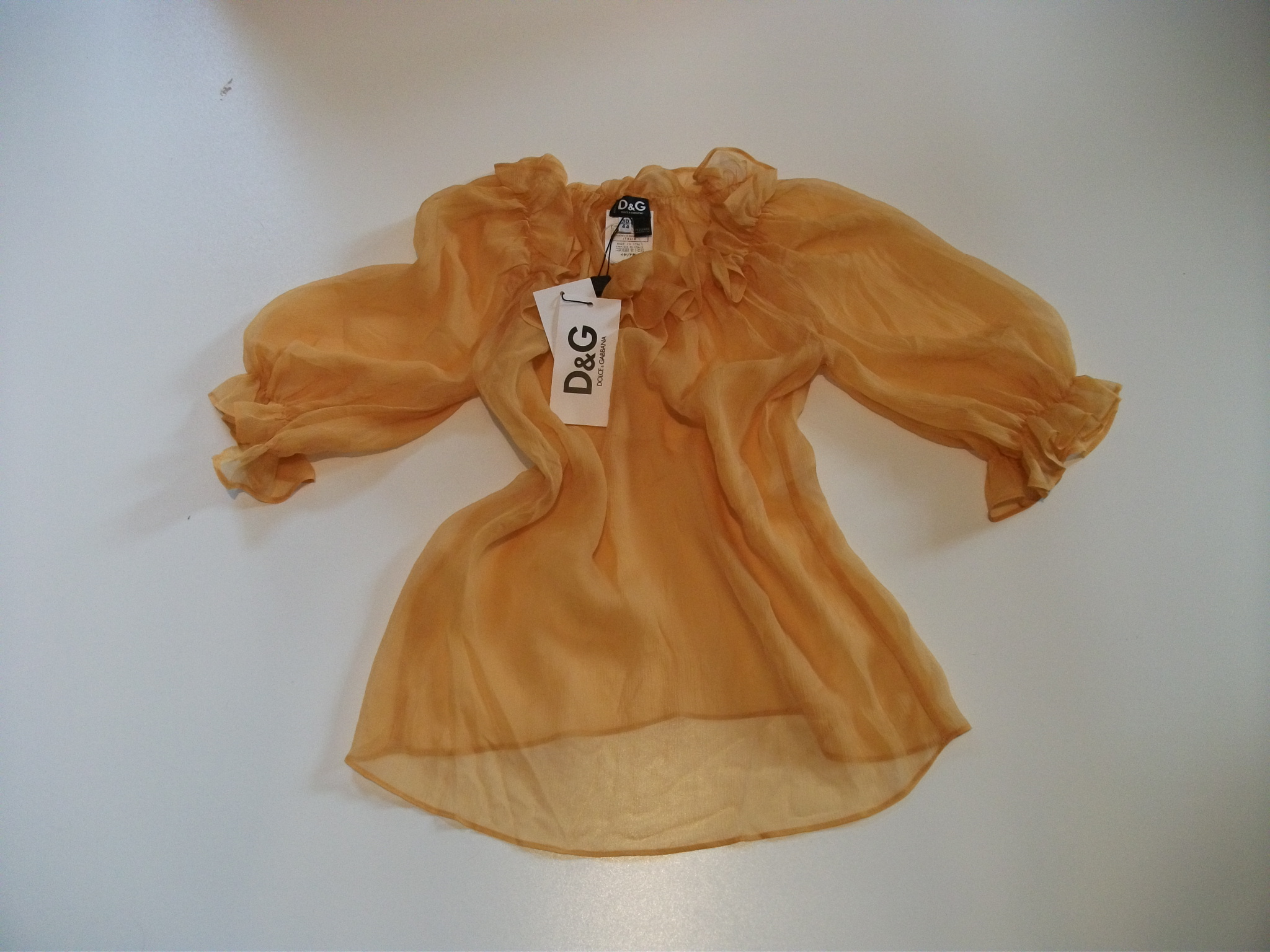 Przezroczysta, pomarańczowa bluzka z długim rękawem i falbanami przy dekolcie, leżąca na białym tle, z widoczną metką D&G.