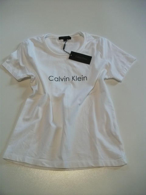 Biała koszulka damska marki Calvin Klein z czarną metką, leżąca na jasnym tle.