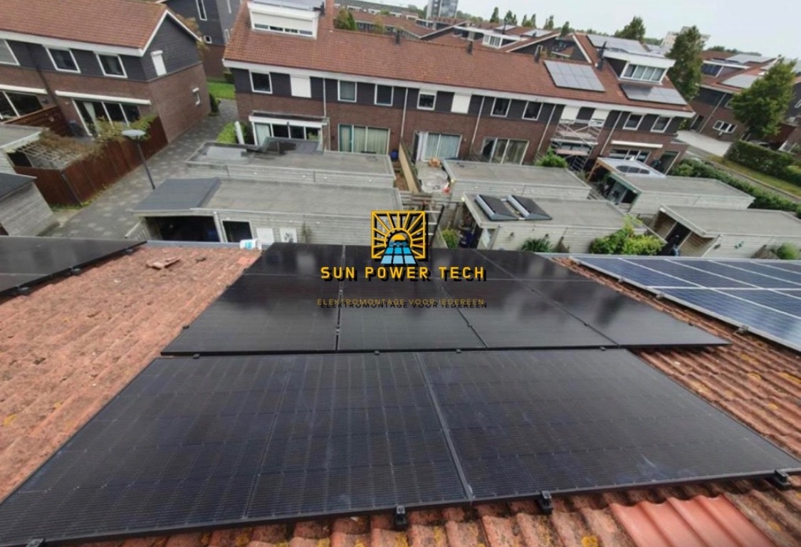 Panele fotowoltaiczne z logo firmy Sun Power Tech zainstalowane na dachu z czerwonej dachówki, widok z góry na osiedle domów jednorodzinnych.