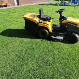 DIRECT BRAND S.C. - Żółta kosiarka traktorowa na idealnie przystrzyżonym trawniku przed brązowym domem z panelami słonecznymi na dachu, w tle plac zabaw dla dzieci.