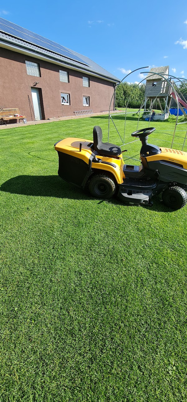 Żółta kosiarka traktorowa na idealnie przystrzyżonym trawniku przed brązowym domem z panelami słonecznymi na dachu, w tle plac zabaw dla dzieci.