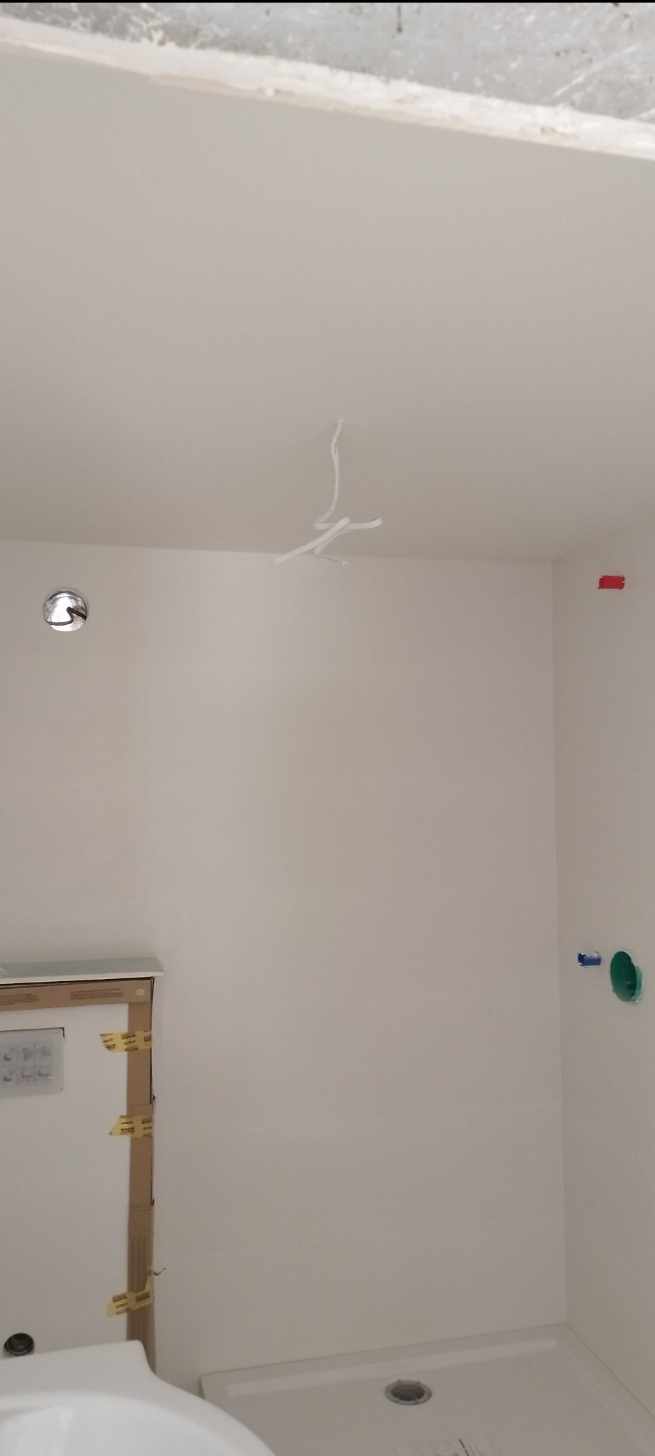 Białe ściany w łazience w trakcie remontu, widoczne wystające kable ze sufitu, przygotowane podejścia wodno-kanalizacyjne i zabezpieczone elementy armatury.