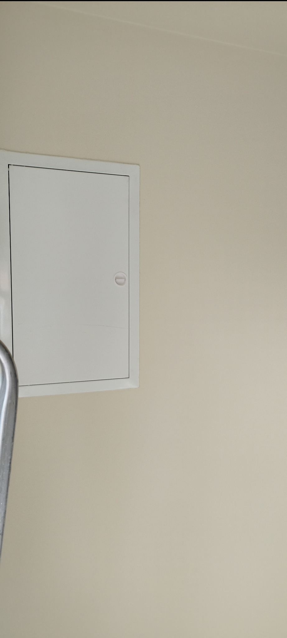Biała klapa rewizyjna na beżowej ścianie, widoczny fragment drabiny malarskiej.