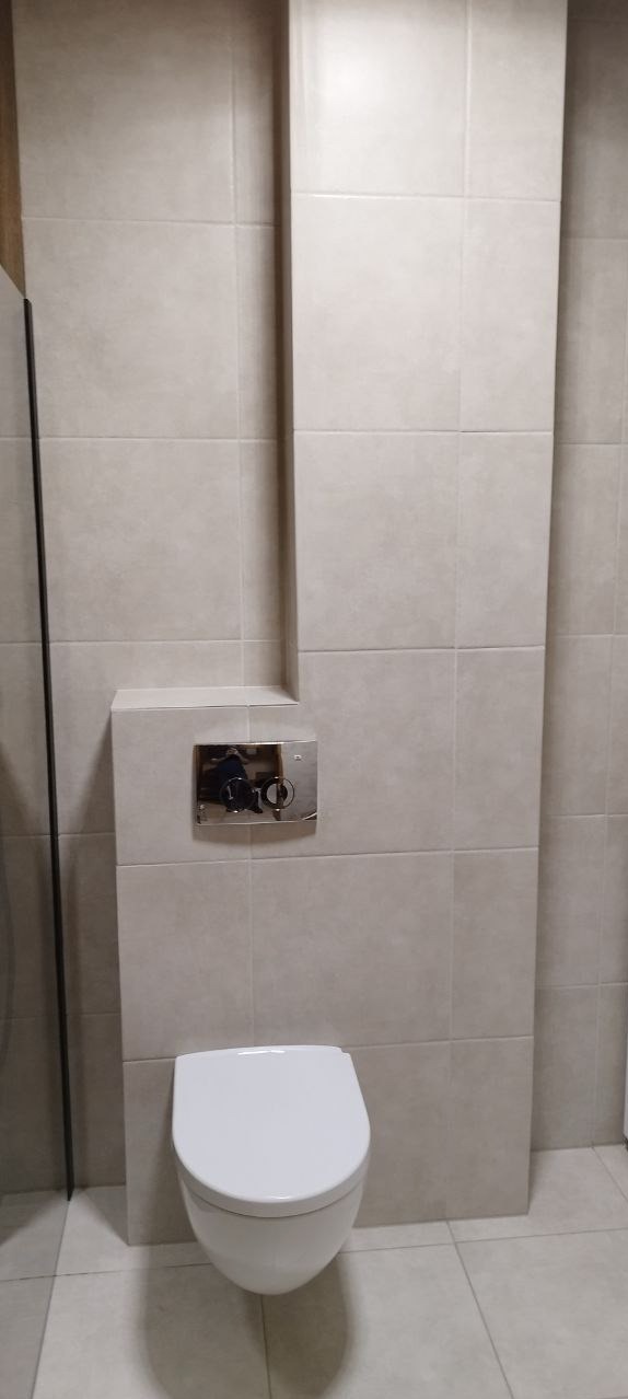 Podwieszana toaleta z chromowanym przyciskiem spłukującym, zamontowana na ścianie wyłożonej jasnymi, kwadratowymi płytkami ceramicznymi.