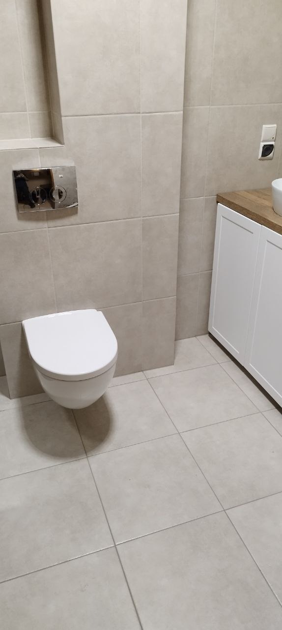 Nowoczesna, podwieszana toaleta w beżowej łazience z płytkami, widoczny panel sterujący spłukiwaniem i biała szafka pod umywalką z drewnianym blatem.