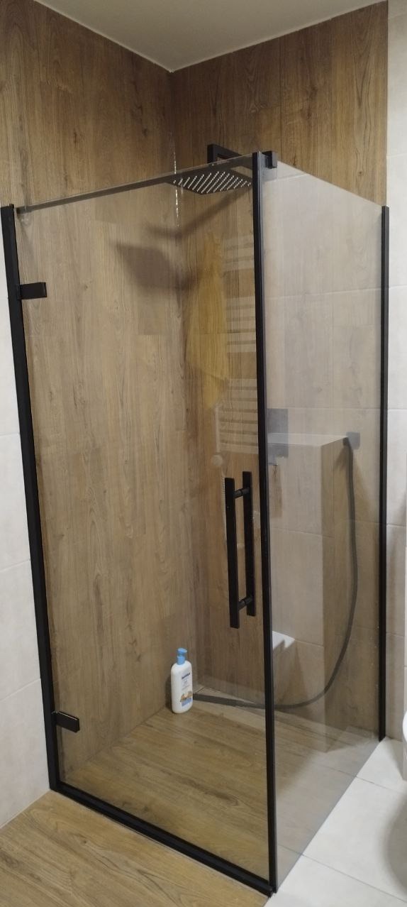 Kabina prysznicowa z czarnymi profilami i deszczownicą w łazience wykończonej płytkami imitującymi drewno i jasnymi płytkami.