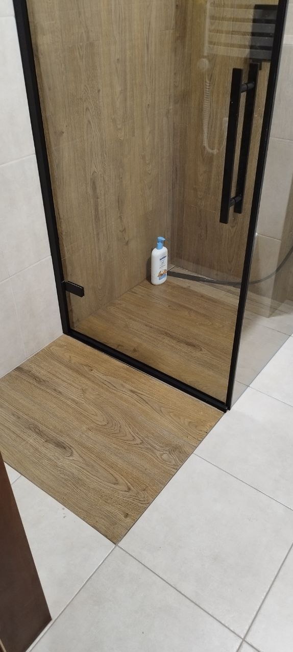 Kabina prysznicowa z drewnopodobną okładziną, czarnymi profilami i uchwytem, na podłodze białe płytki, w rogu butelka kosmetyku.