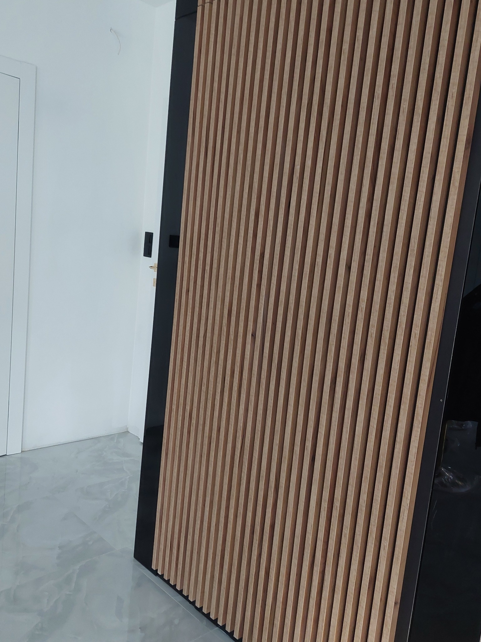 Nowoczesny panel ścienny z pionowych, drewnianych listew w jasnym odcieniu, obramowany czarną ramą, na tle białej ściany i szarej podłogi z marmurowym wzorem.