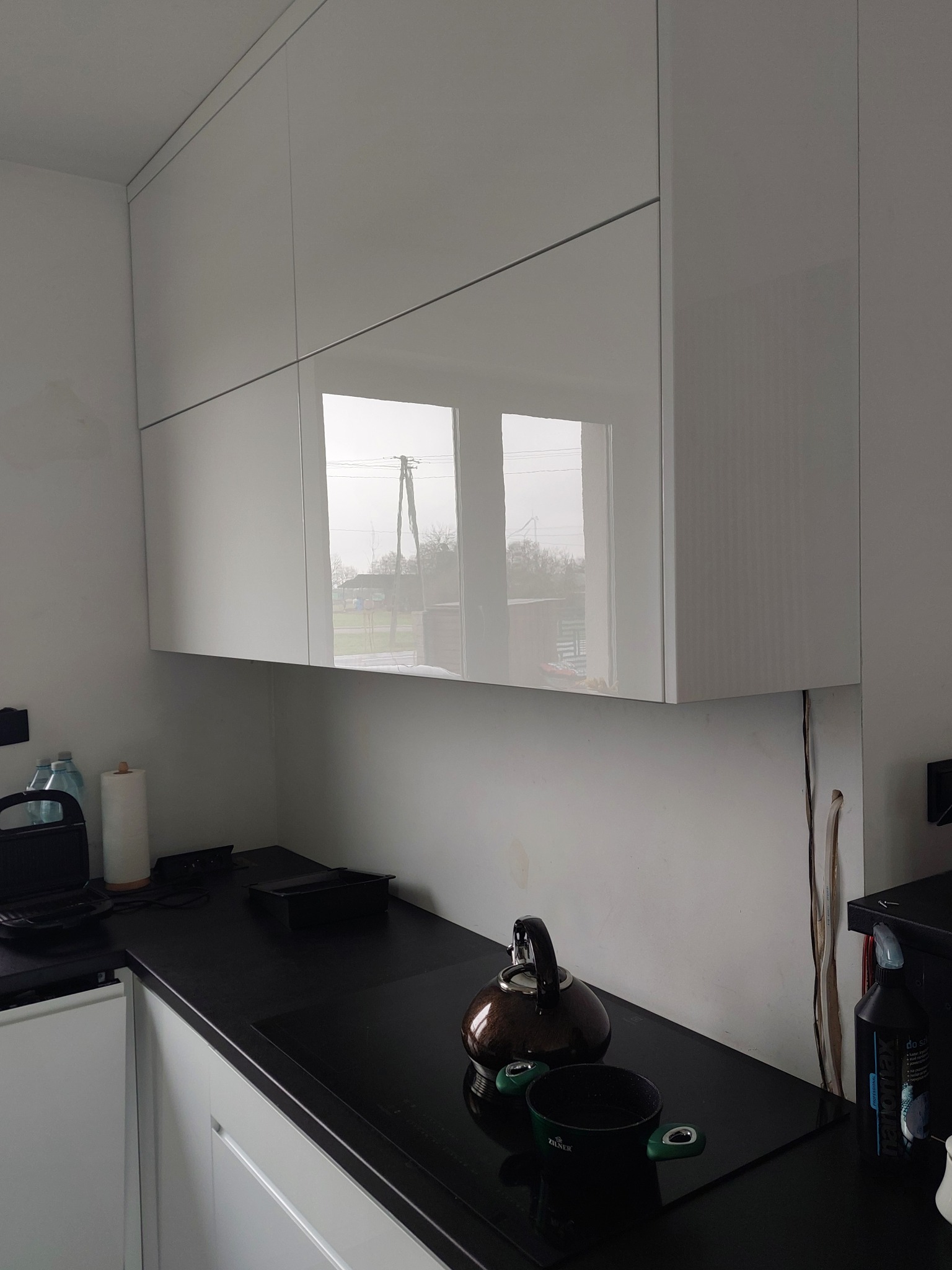 Białe, lakierowane szafki kuchenne wiszące, z odbiciem okna w frontach, czarny blat kuchenny z czajnikiem i garnkiem.