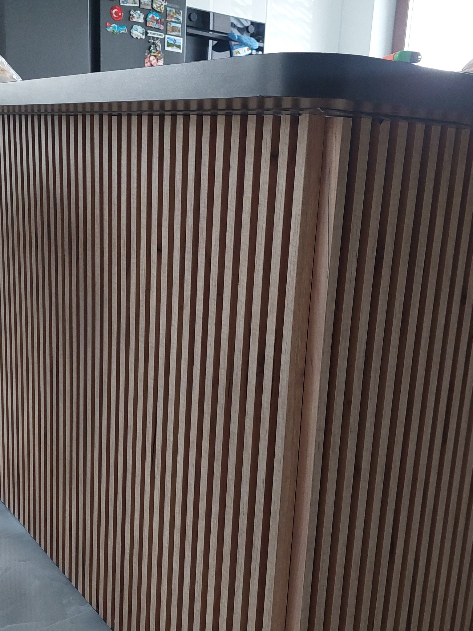 Fragment baru kuchennego z pionowymi, drewnianymi lamelami w odcieniach brązu, wykończonego czarnym blatem, widoczny fragment szarej podłogi i lodówki z magnesami.