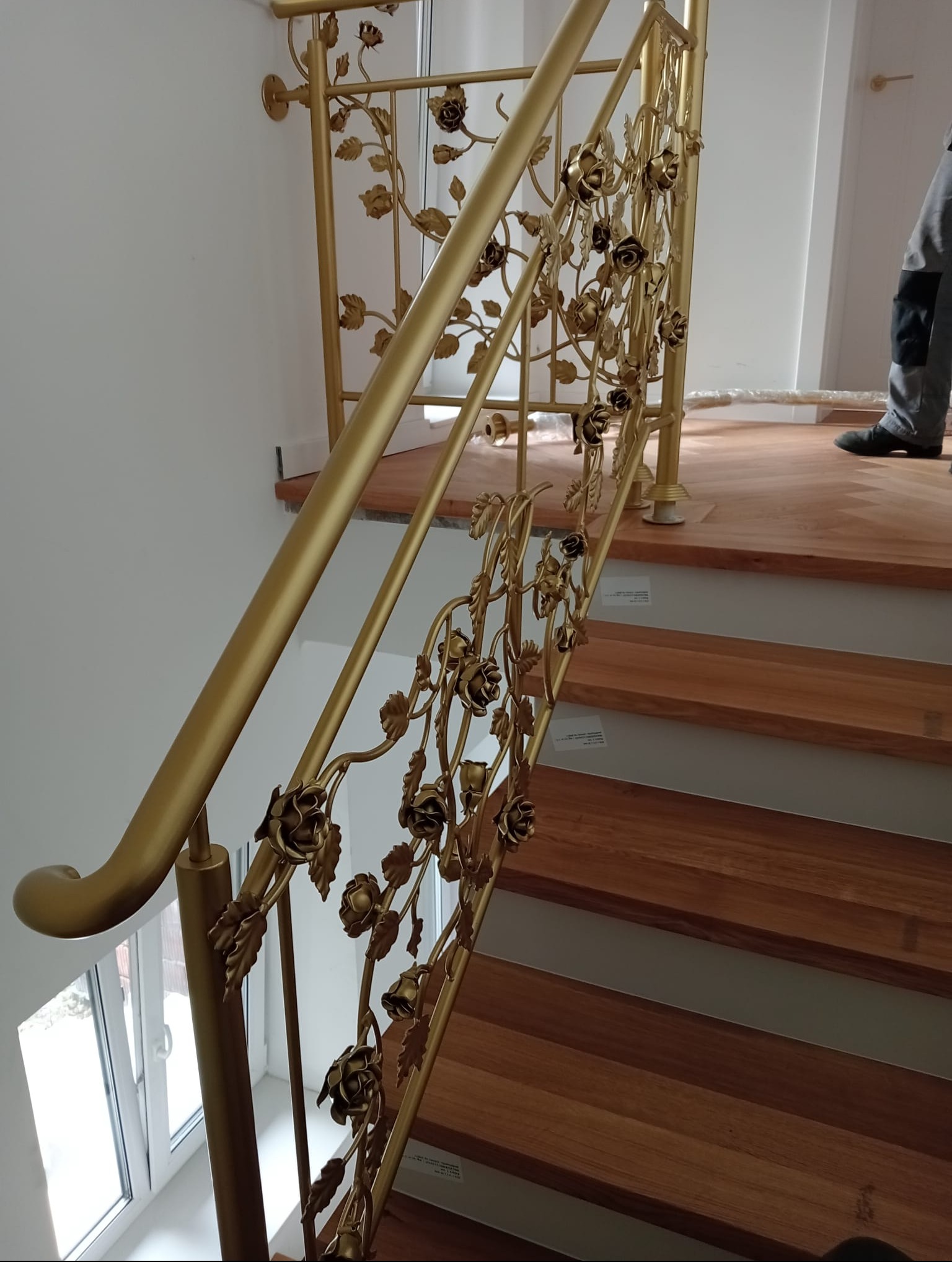 Złota balustrada schodowa z motywem kwiatowym róży, zamontowana na drewnianych schodach; widoczny fragment osoby w tle.