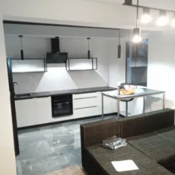 Nowoczesna kuchnia z białymi szafkami i czarnymi akcentami, widok z salonu, podłoga z szarego gresu polerowanego, oświetlenie punktowe i wiszące, sofa w odcieniach szarości w pierwszym planie.