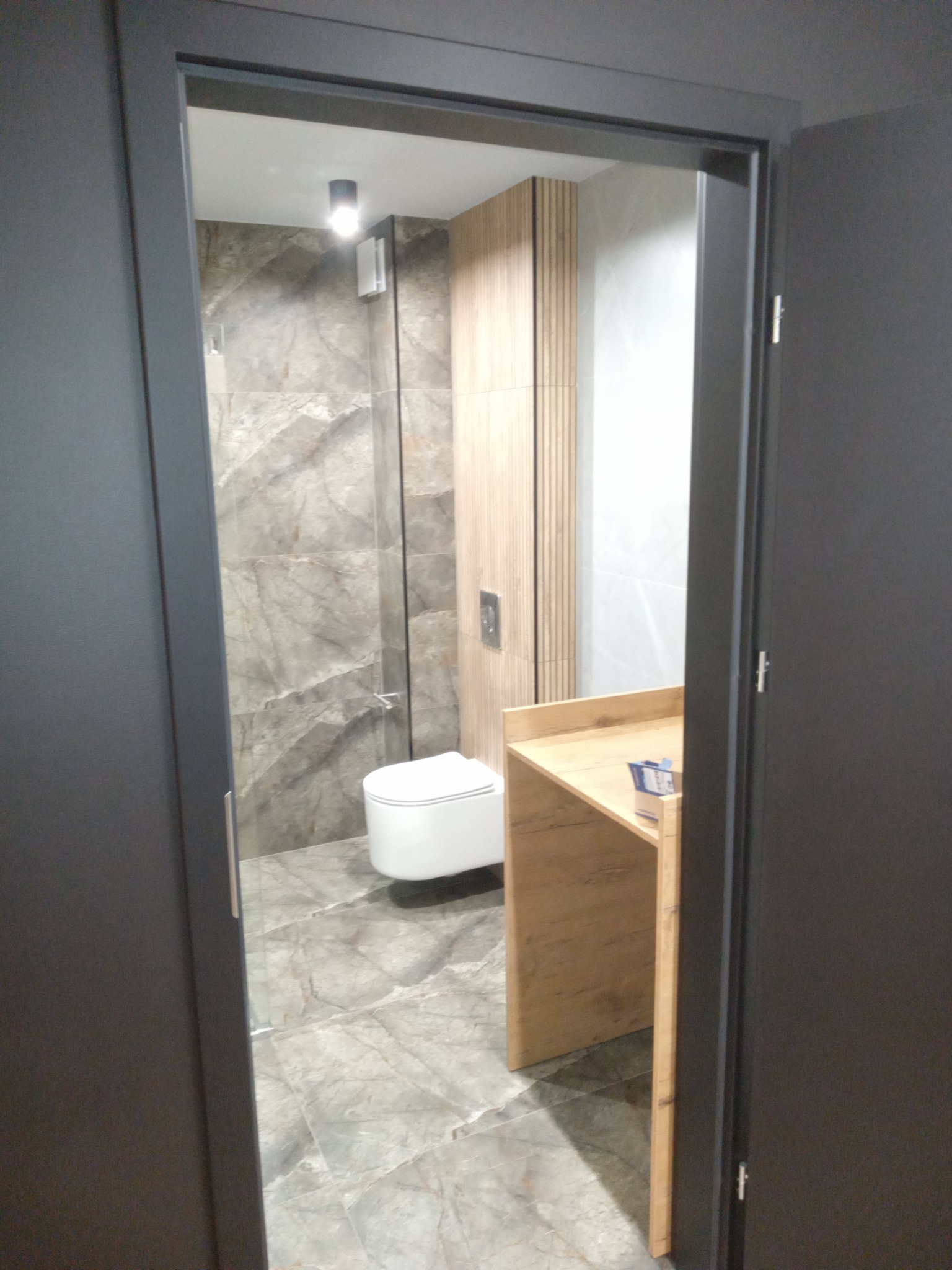 Widok przez otwarte, ciemne drzwi na wykończoną łazienkę z marmurowymi płytkami, podwieszaną toaletą i drewnianą szafką z blatem.