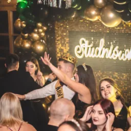 Uczestnicy studniówki w trakcie zabawy, widoczny napis 'Studniówka' z dekoracjami balonowymi w tle. Młodzież w strojach wieczorowych.