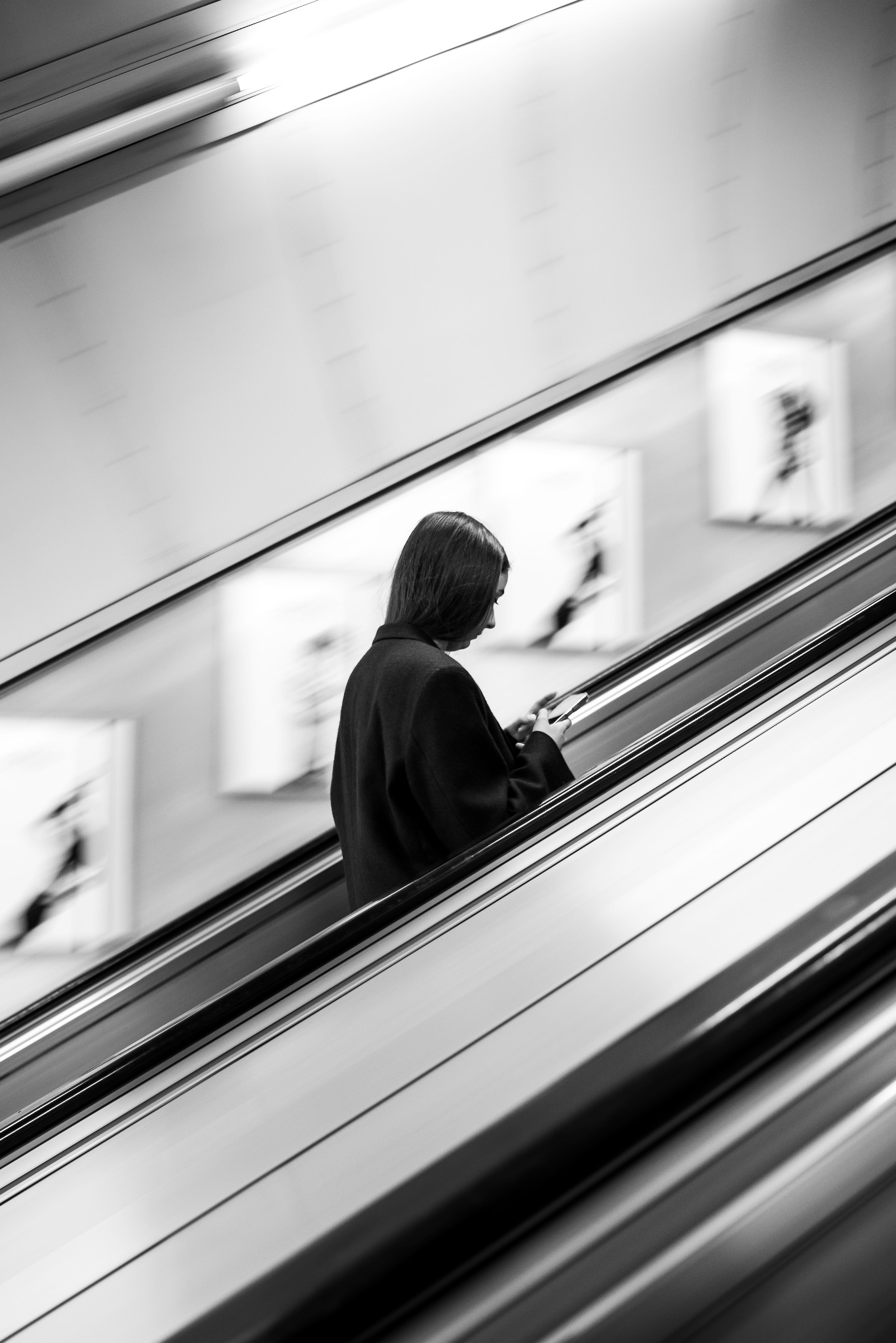 Monochromatyczne ujęcie kobiety na ruchomych schodach, pochylonej nad telefonem. W tle rozmyte plakaty reklamowe.