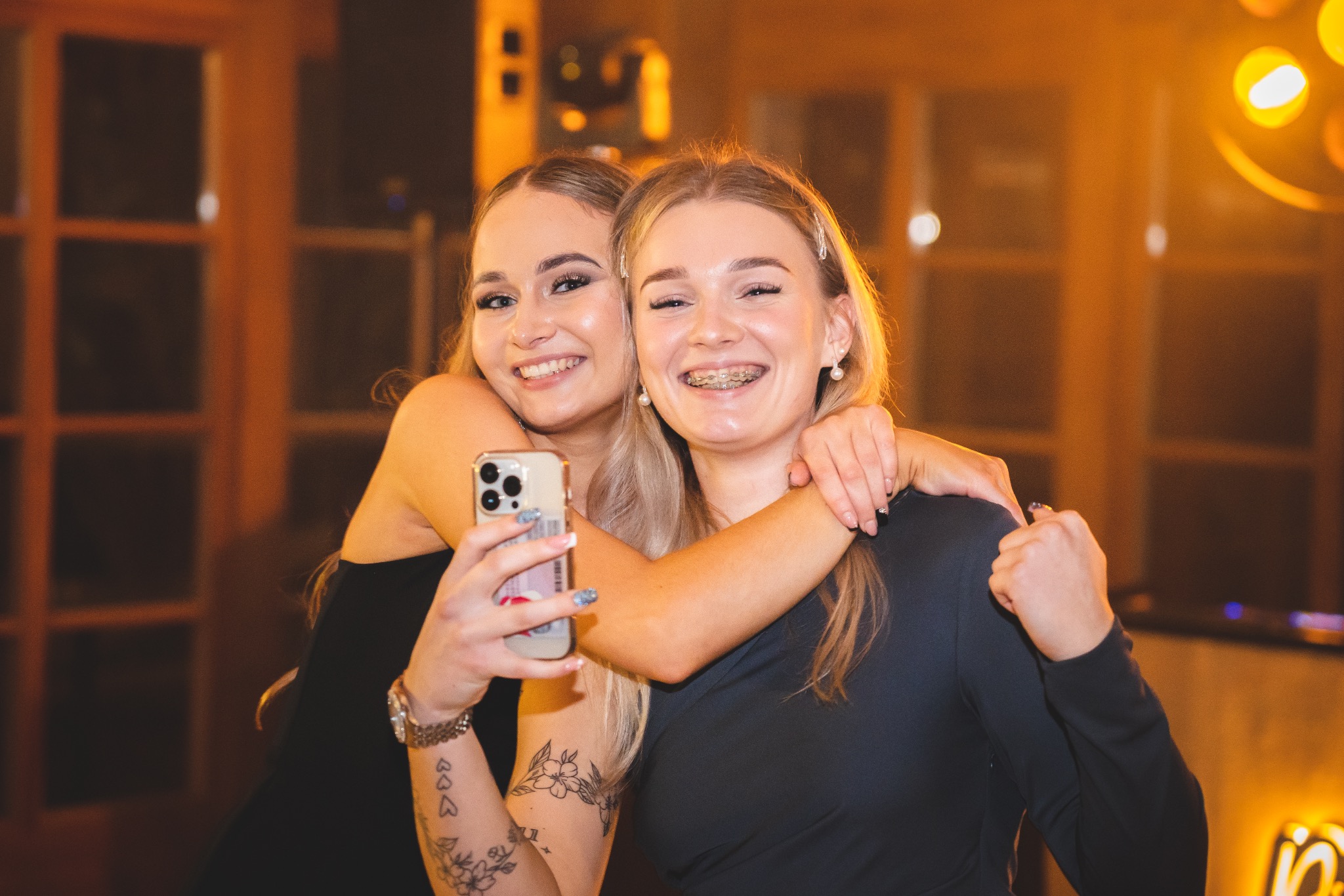 Dwie uśmiechnięte nastolatki obejmują się, robiąc selfie telefonem iPhone w ciepłym oświetleniu wnętrza. Jedna z dziewcząt ma aparat ortodontyczny.