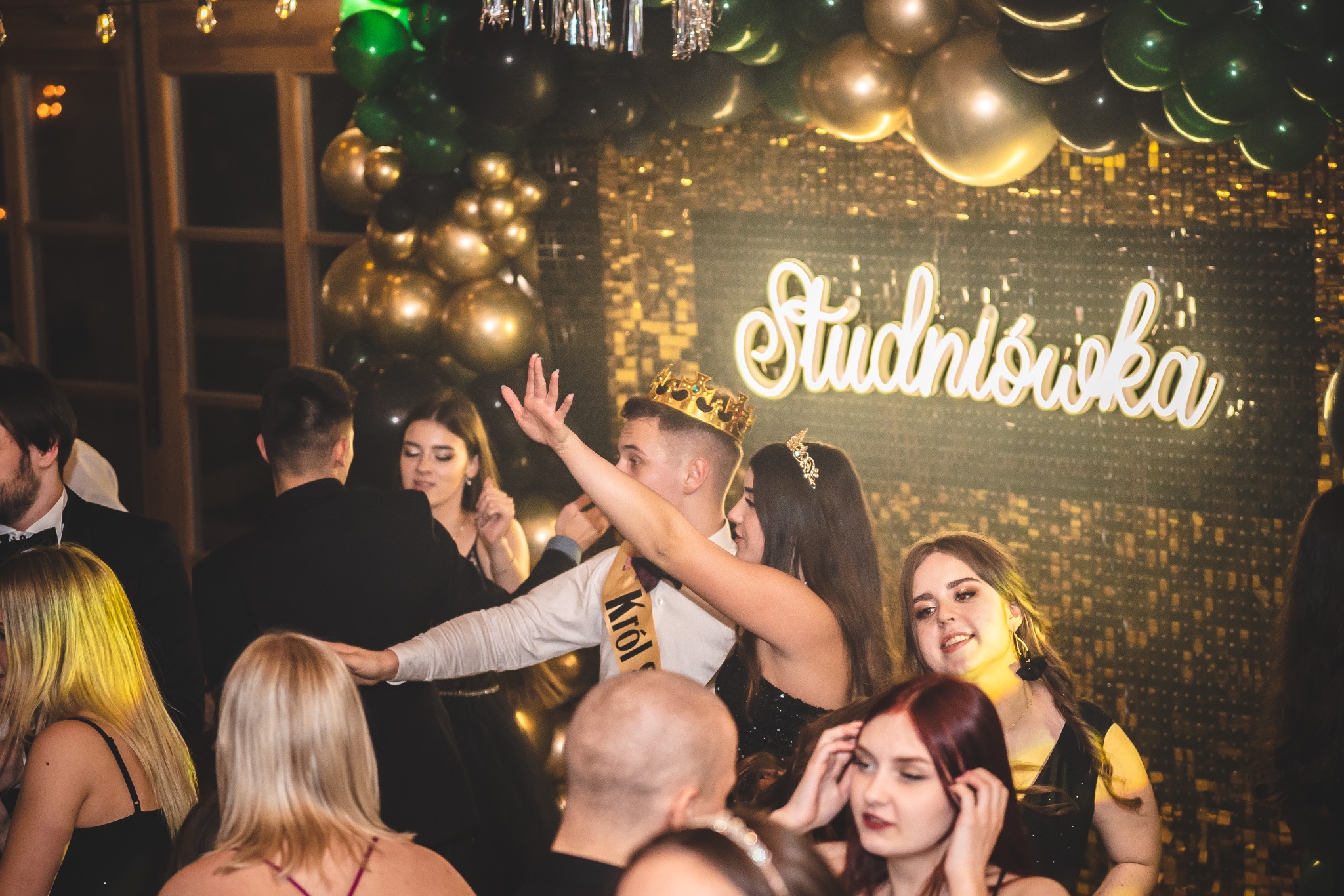 Uczestnicy studniówki w trakcie zabawy, widoczny napis 'Studniówka' z dekoracjami balonowymi w tle. Młodzież w strojach wieczorowych.
