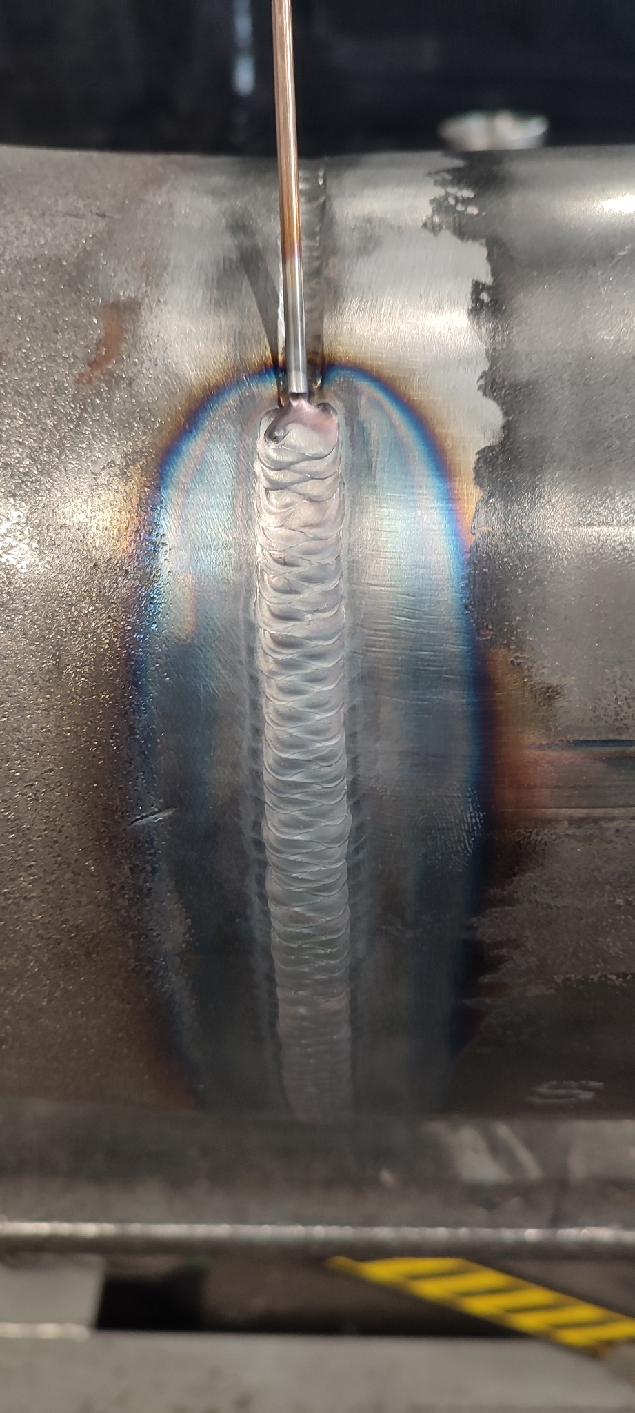 Świeżo wykonany spaw na metalowej rurze, widoczny tęczowy efekt termiczny wokół spoiny. Precyzyjne łączenie metali, zbliżenie na detal spawalniczy.