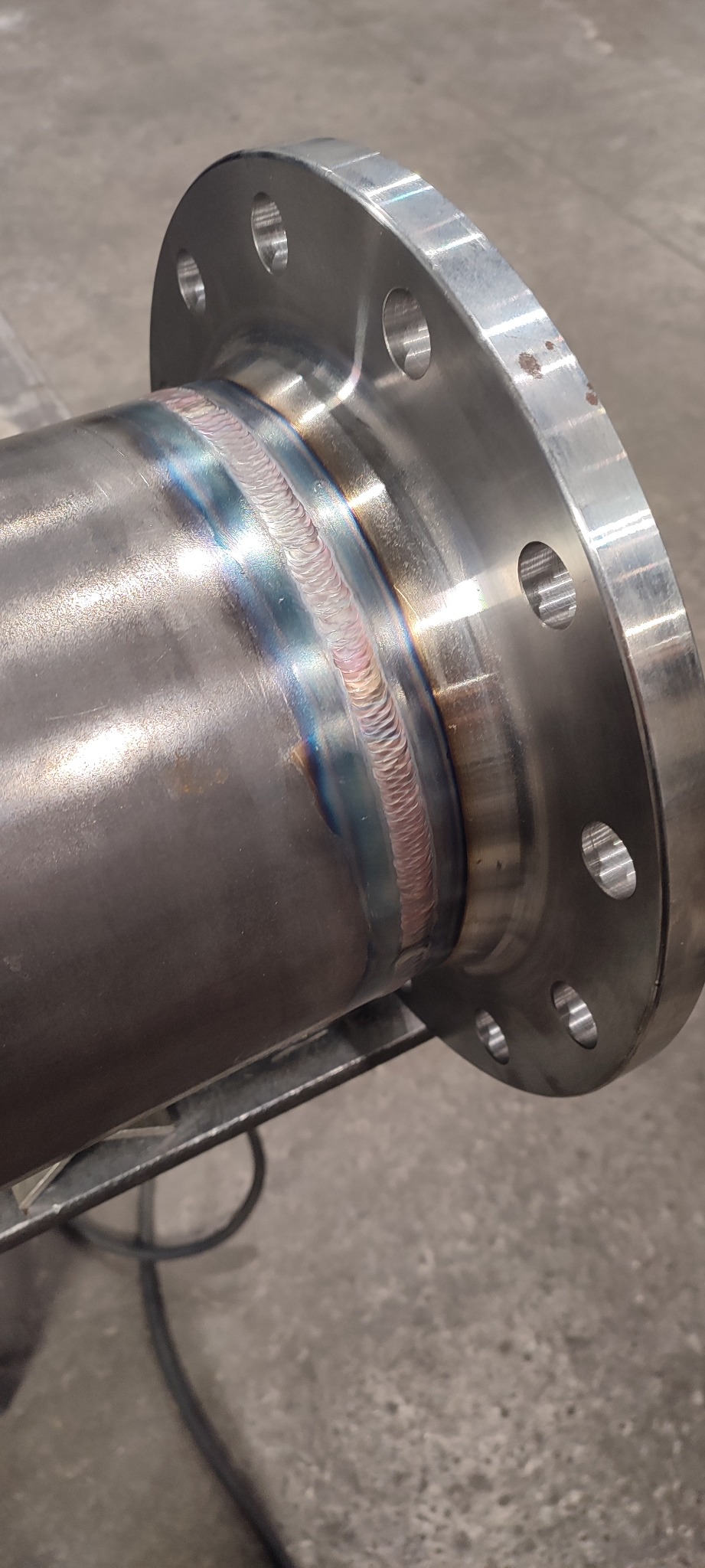 Precyzyjny spaw rury ze stalową flanszą, widoczne tęczowe przebarwienia po spawaniu, świadczące o wysokiej temperaturze i jakości wykonania.