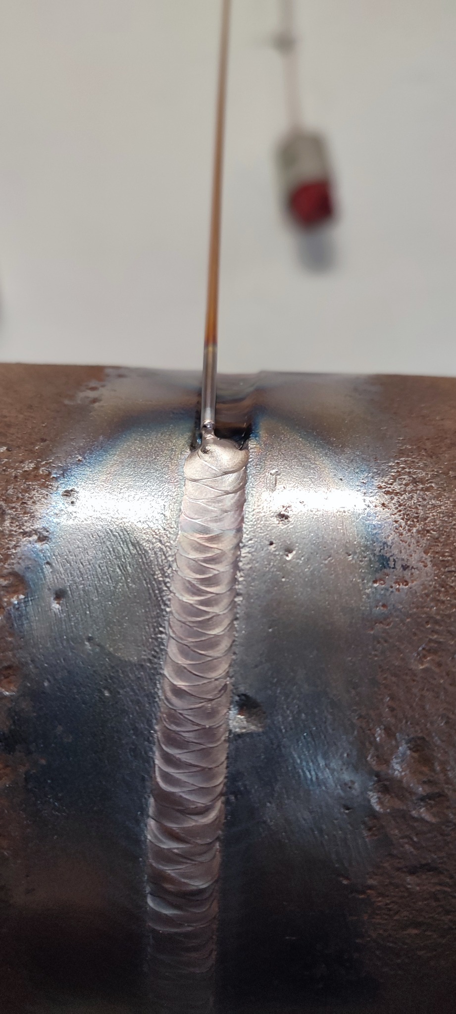 Detal spoiny na metalowej rurze, wykonanej metodą TIG. Widoczne tęczowe przebarwienia termiczne i precyzyjne ułożenie ściegu. W tle biała ściana i uchwyt spawalniczy.