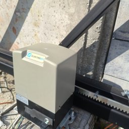 Pat-instal - Szary automat do bram przesuwnych Elektrobim zamontowany na betonowej podstawie, widoczna listwa zębata i przewody.
