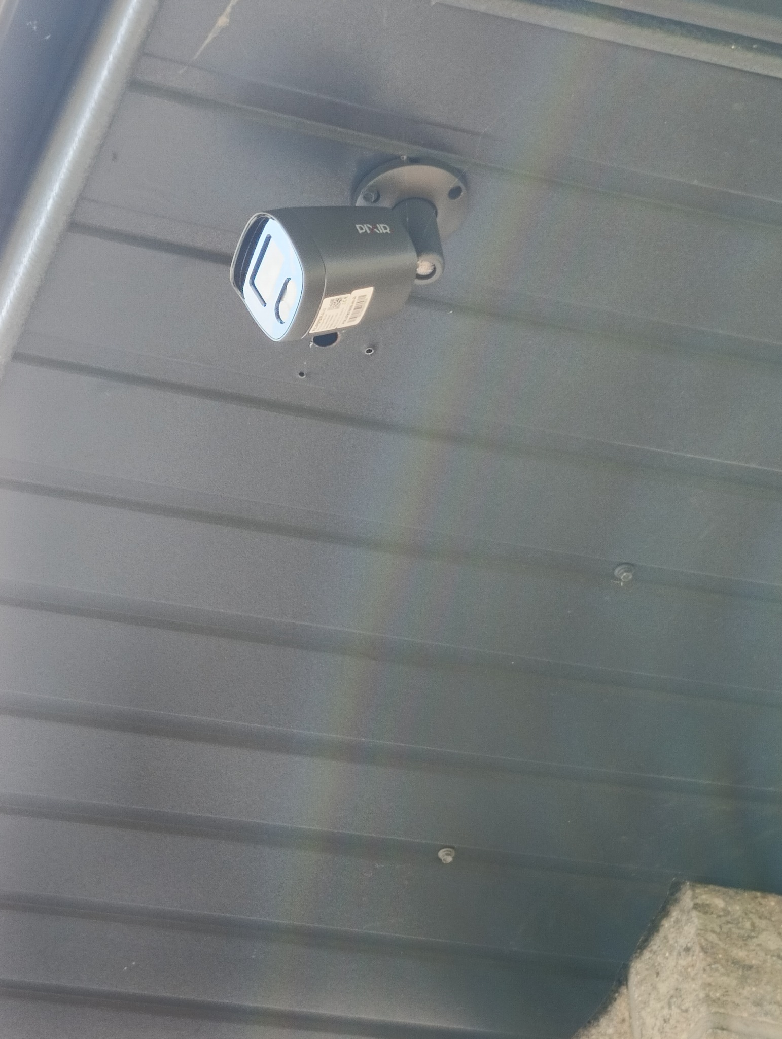Czarna kamera monitoringu marki Pixir zamontowana na szarej, metalowej powierzchni z widocznymi śrubami mocującymi i delikatną tęczą światła.