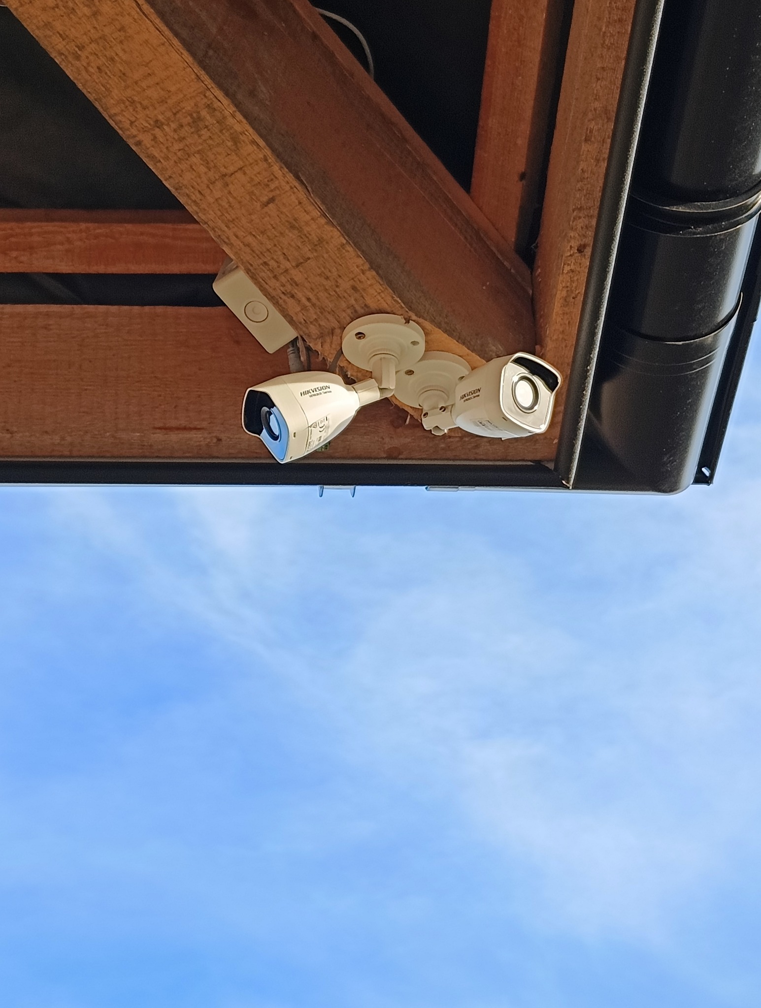 Dwie białe kamery monitoringu Hikvision zamontowane pod drewnianym okapem, widok z dołu na błękitne niebo.