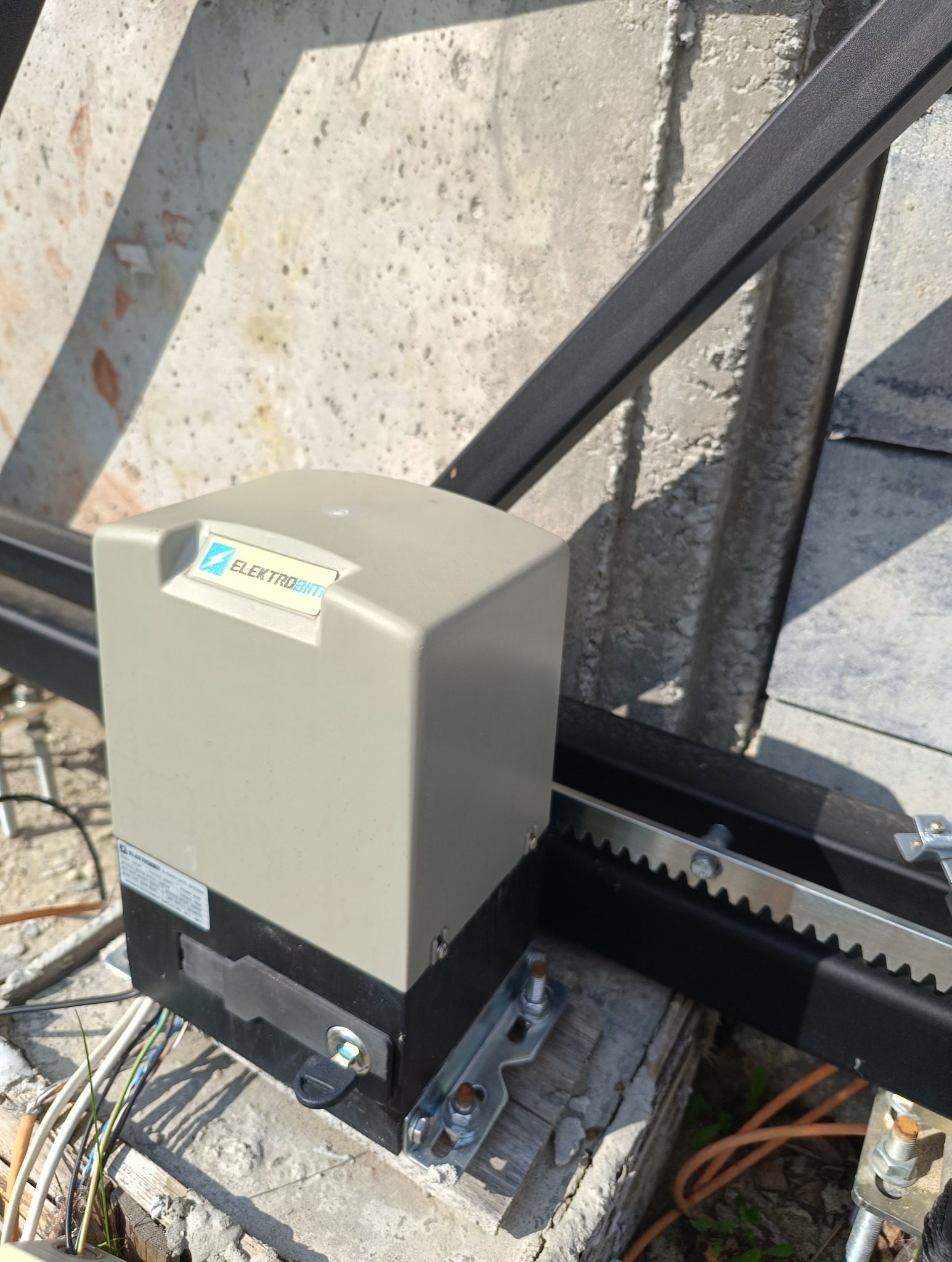 Szary automat do bram przesuwnych Elektrobim zamontowany na betonowej podstawie, widoczna listwa zębata i przewody.
