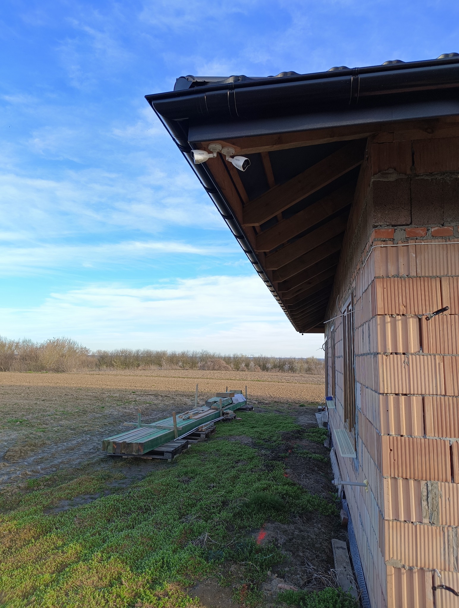 Dwie kamery monitoringu zamontowane pod okapem surowego budynku z czerwonej cegły kratówki, w tle pole i błękitne niebo.