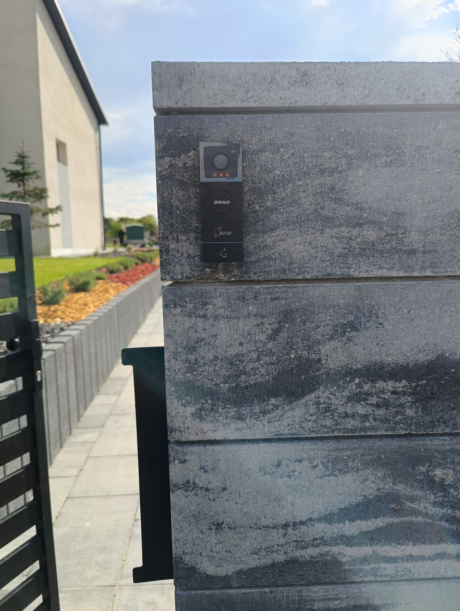 Szary, kamienny słupek bramy z zamontowanym wideodomofonem ORNO z czytnikiem RFID i przyciskiem dzwonka, widok z boku, w tle fragment domu i zagospodarowany ogród.