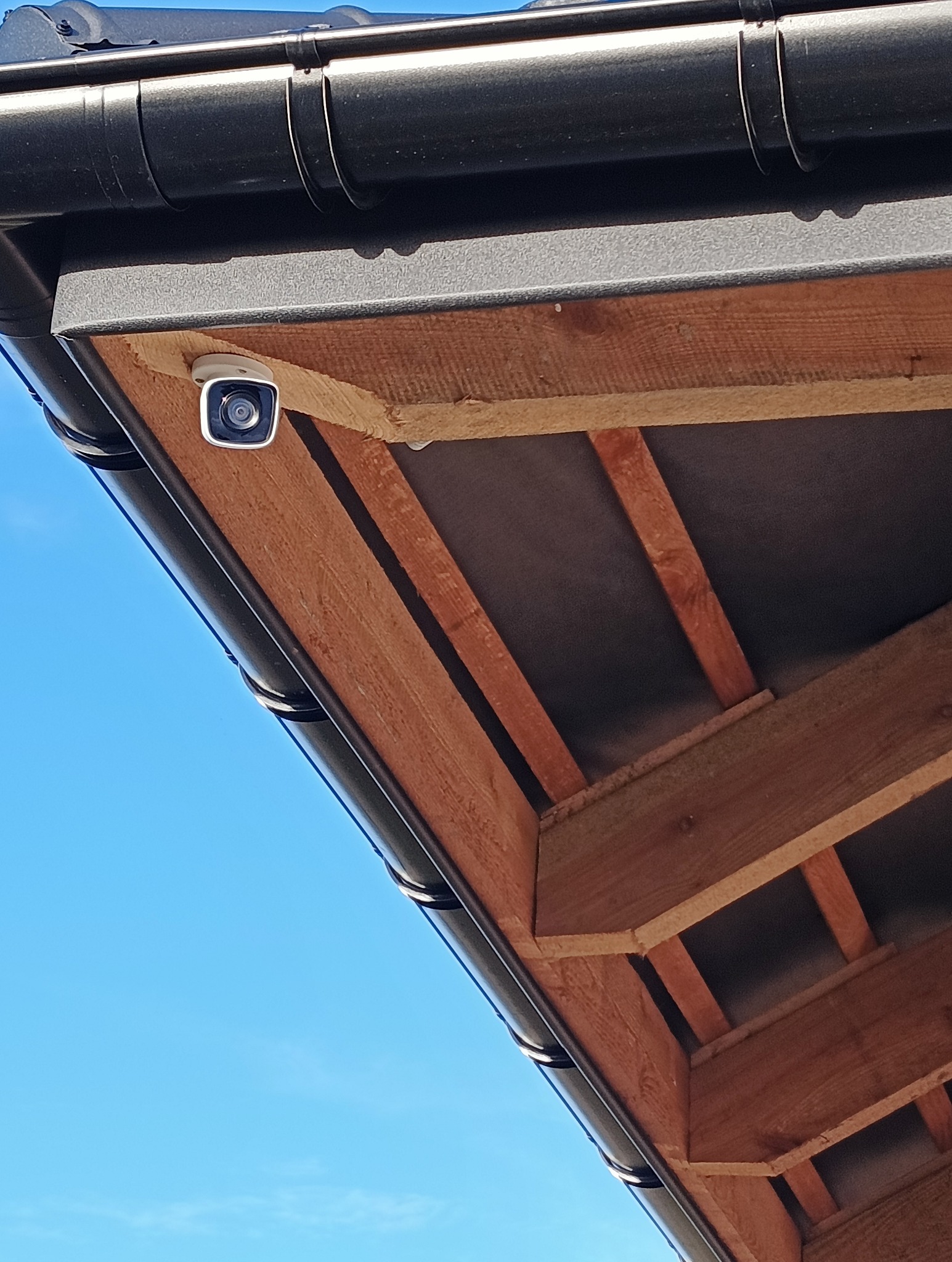 Kamera monitoringu zamontowana pod okapem dachu, widoczna drewniana konstrukcja dachu i fragment błękitnego nieba.