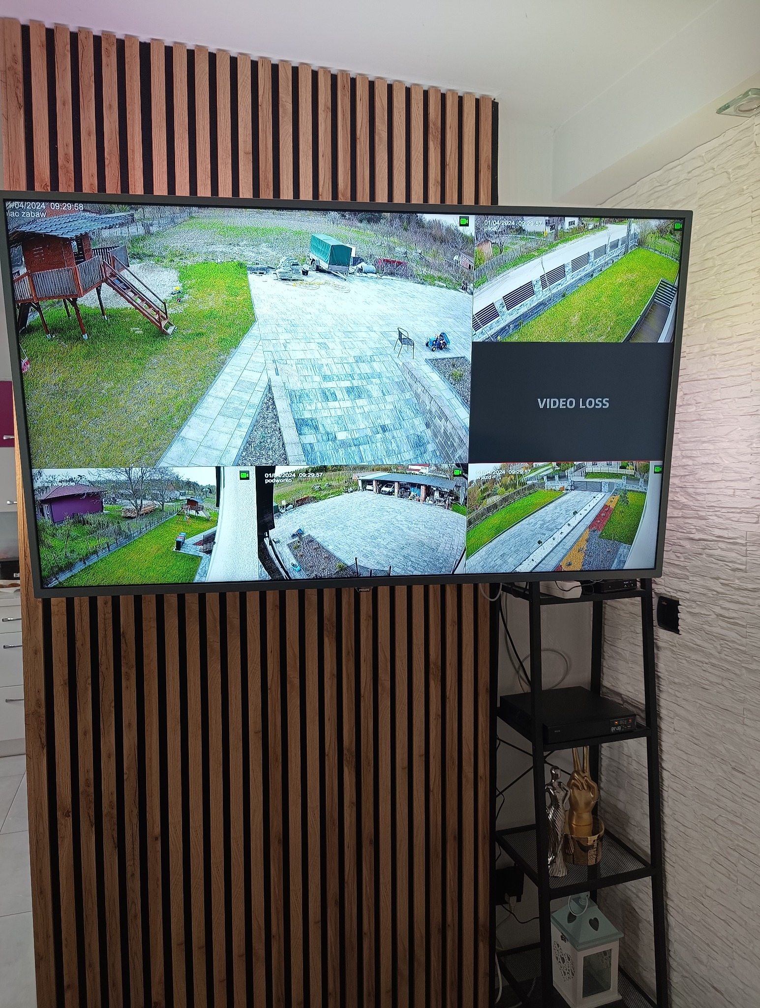 Ekran telewizora prezentujący obraz z systemu kamer monitoringu obejmującego różne obszary posesji, w tym podwórko, ogród i wjazd.