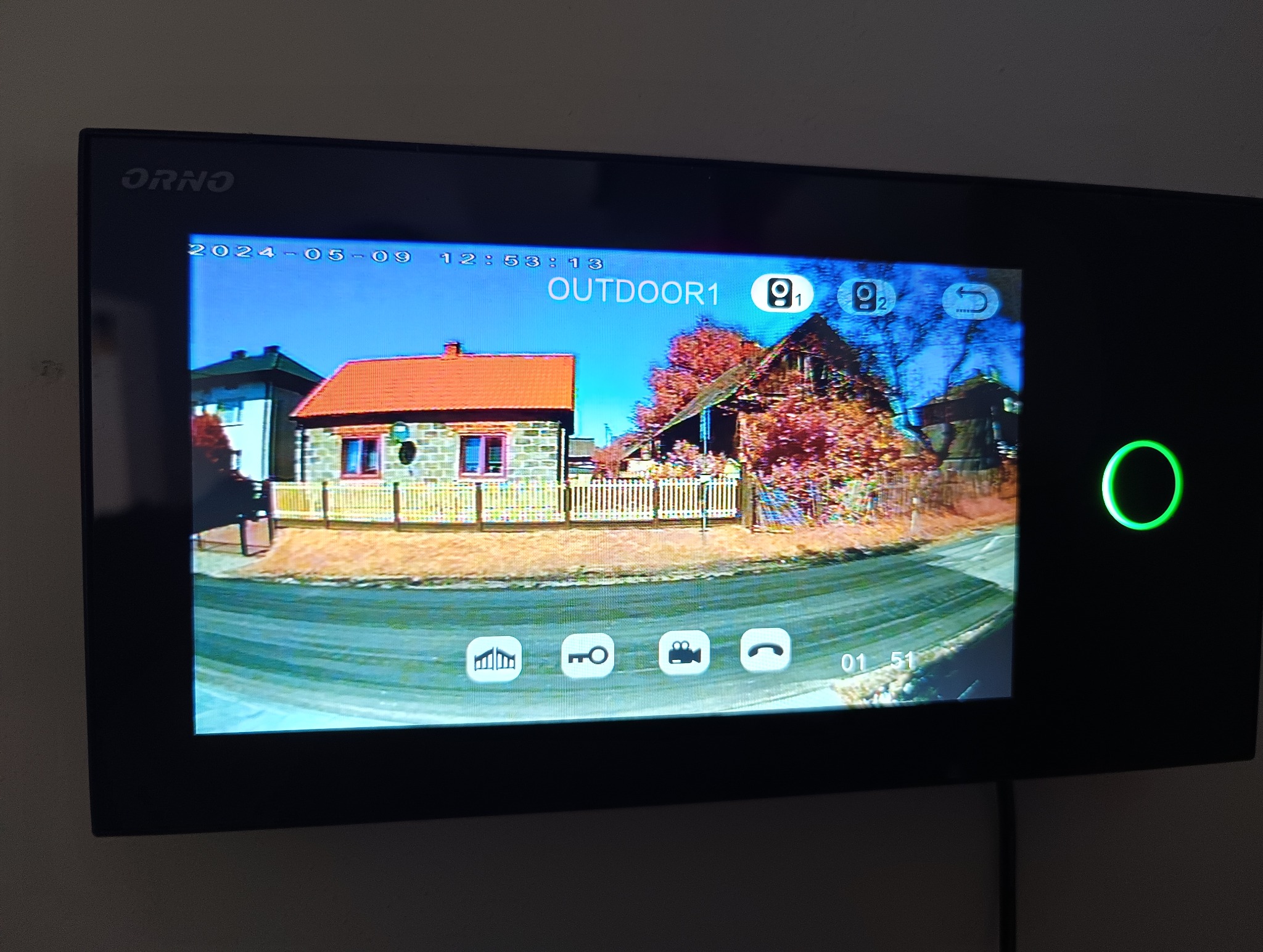 Ekran wideodomofonu ORNO z widokiem kamery na dom z czerwonym dachem i drewnianym płotem, data i godzina w lewym górnym rogu, ikony sterowania na dole.