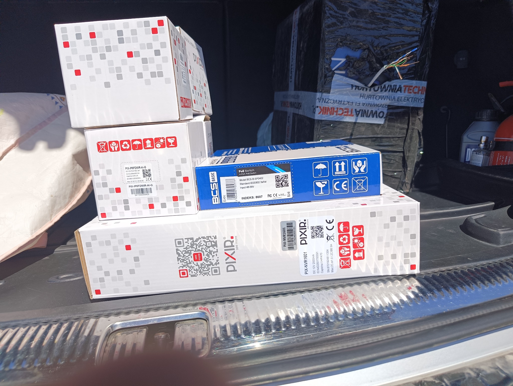 Paczki z urządzeniami do monitoringu i alarmów, w tym switchem PoE, leżące w bagażniku samochodu, widoczne kable i akcesoria montażowe.