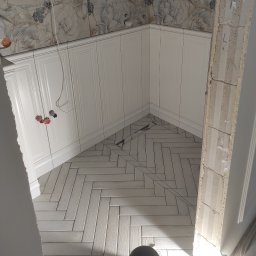 Oleksandr Ryzhenko - Białe szafki kuchenne z frezowanymi frontami i ułożona w jodełkę podłoga z płytek imitujących drewno, widoczne fragmenty instalacji wodnej i fragment ściany z dekoracyjną tapetą w kwiaty podczas...