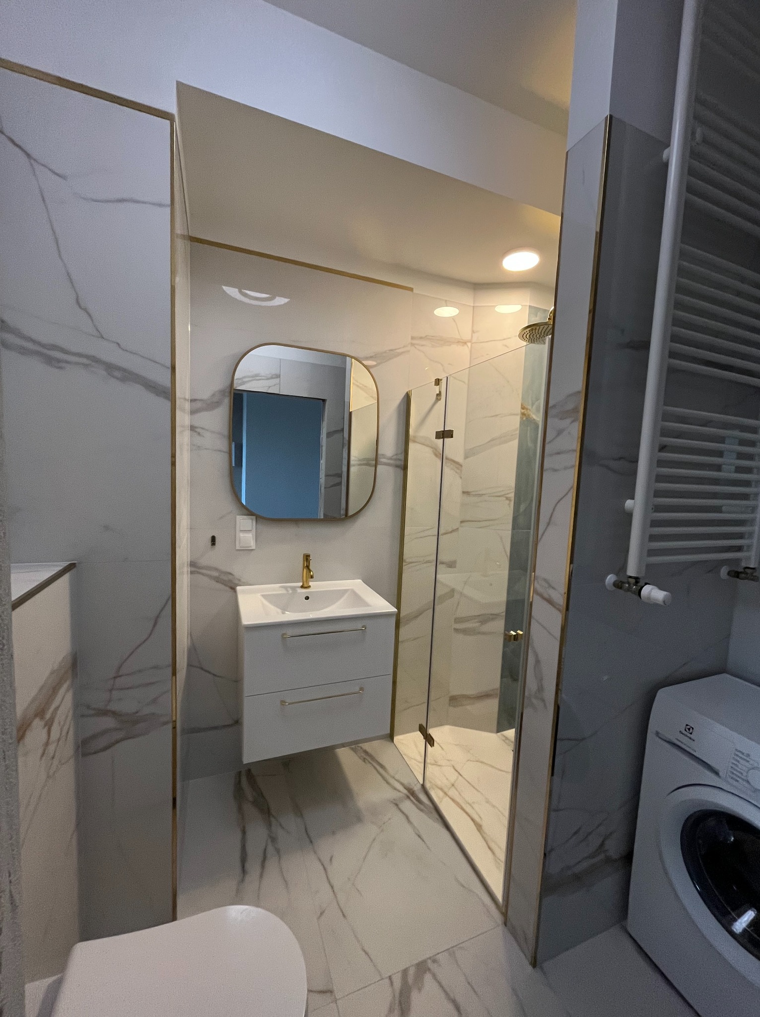 Nowoczesna łazienka w bieli i marmurze, z kabiną prysznicową ze złotymi detalami, umywalką z szafką, okrągłym lustrem i pralką Electrolux.
