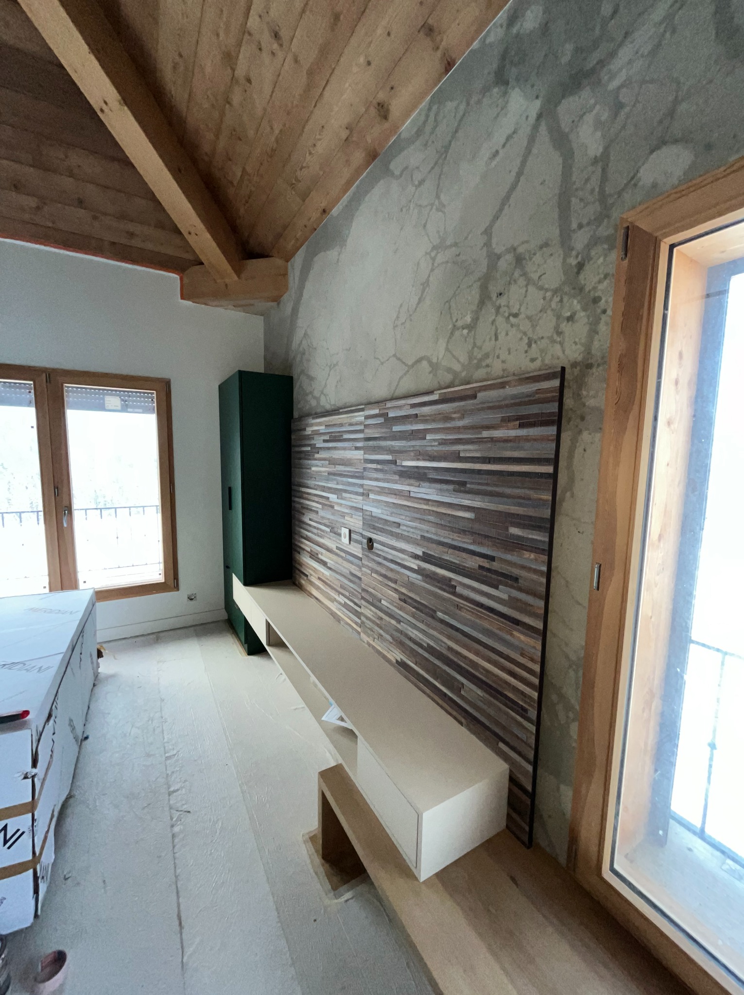 Wnętrze w trakcie wykańczania: drewniany sufit, ściana z efektem betonu, panel dekoracyjny z drewnianych listew, zielona szafa i biała szafka RTV.