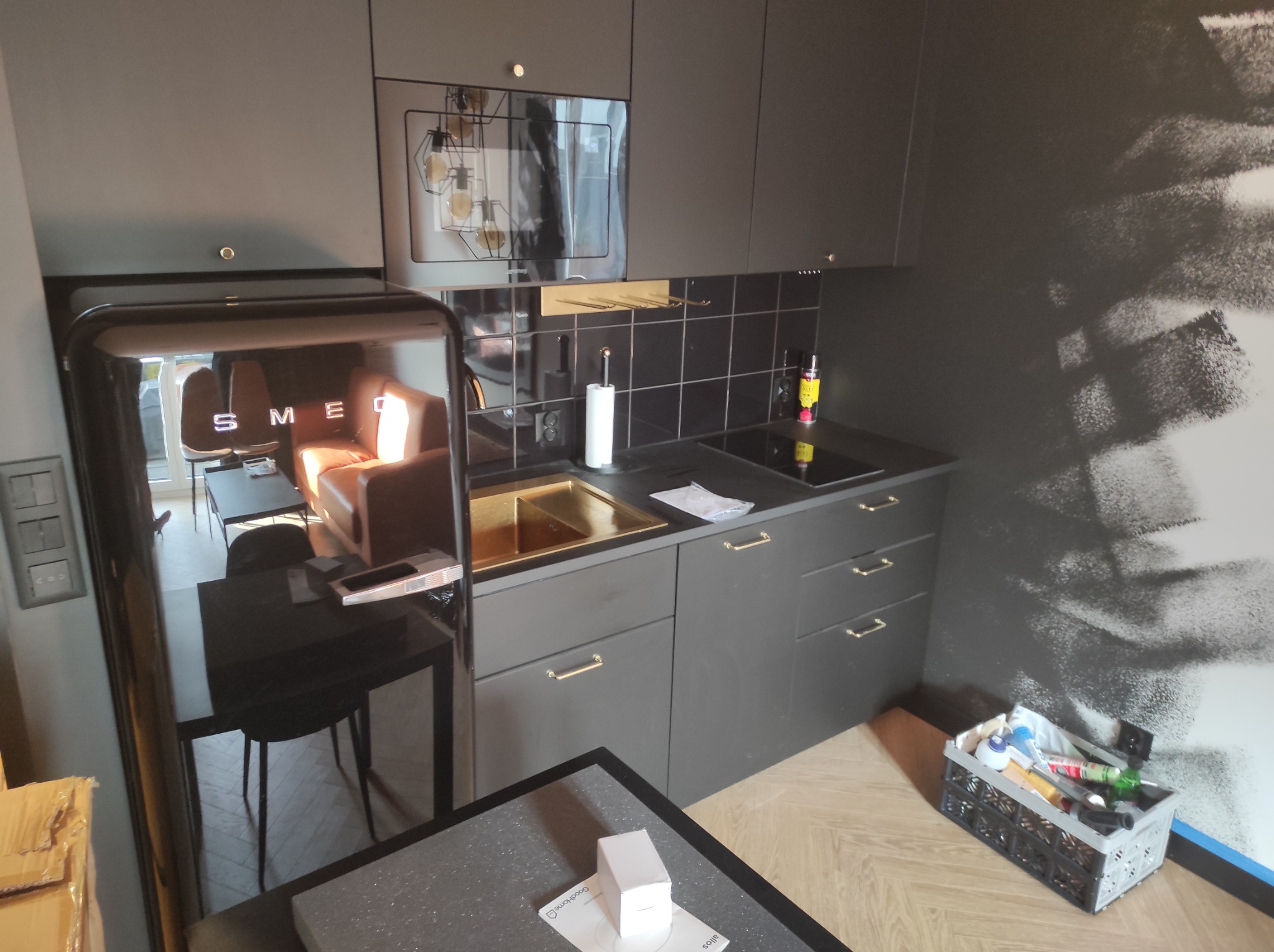 Nowoczesna, szara kuchnia z czarną armaturą i lodówką Smeg, widoczny fragment ściany z abstrakcyjnym wzorem, skrzynka z narzędziami w rogu.