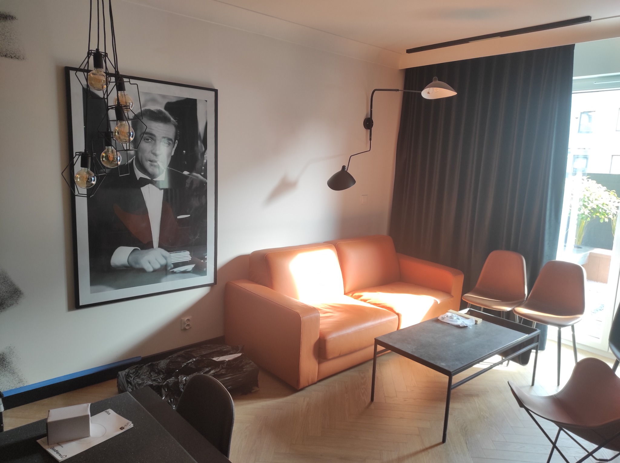 Jasny salon z parkietem, pomarańczową sofą, stolikiem kawowym, krzesłami w stylu retro i plakatem Seana Connery'ego w czarnej ramie.