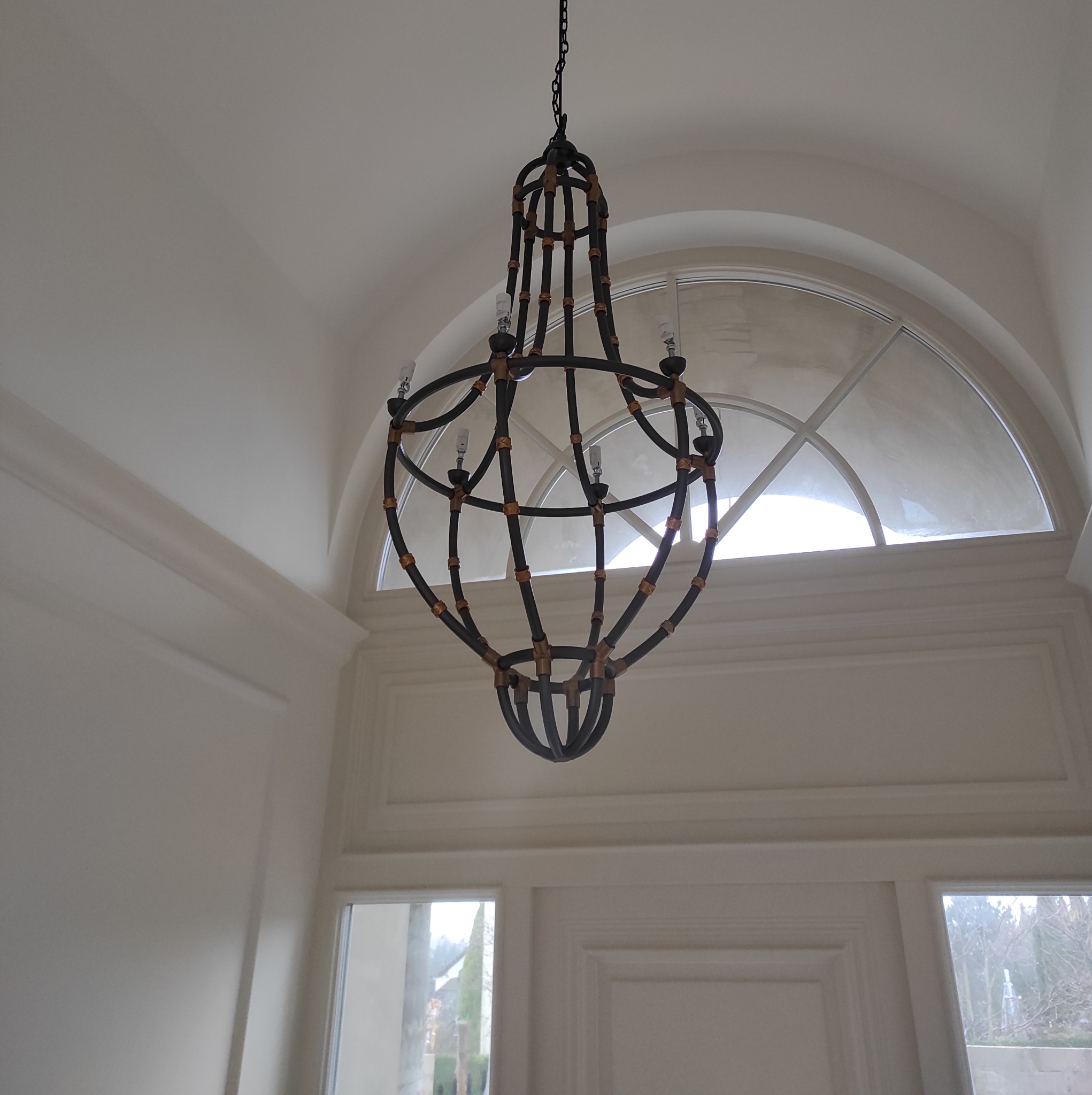 Żyrandol z metalową konstrukcją i drewnianymi akcentami, zawieszony na łańcuchu w jasnym wnętrzu z dużym, łukowatym oknem i sztukaterią.