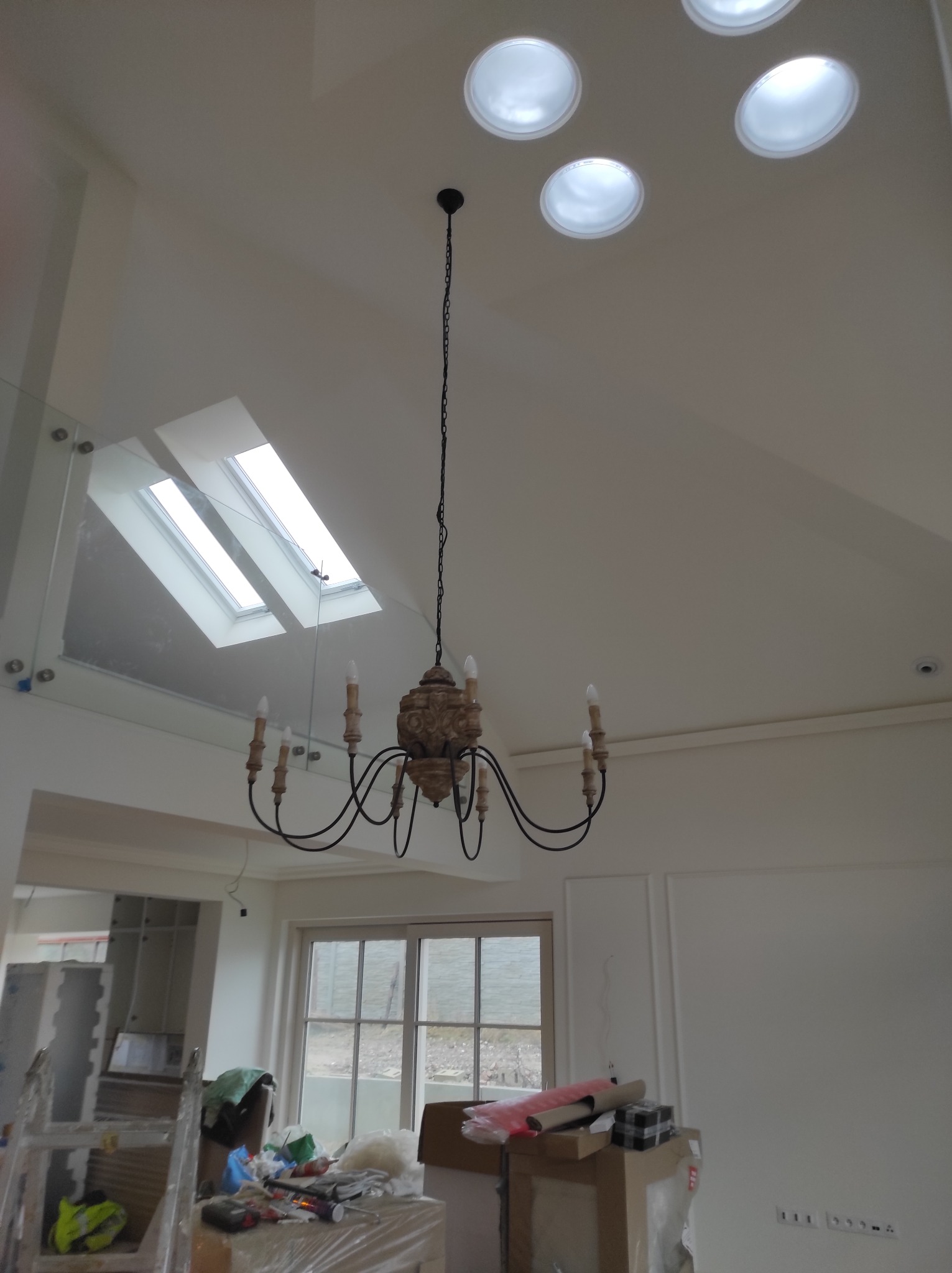 Wnętrze w trakcie prac wykończeniowych: widoczny żyrandol, okna dachowe, oraz materiały budowlane i narzędzia na podłodze.