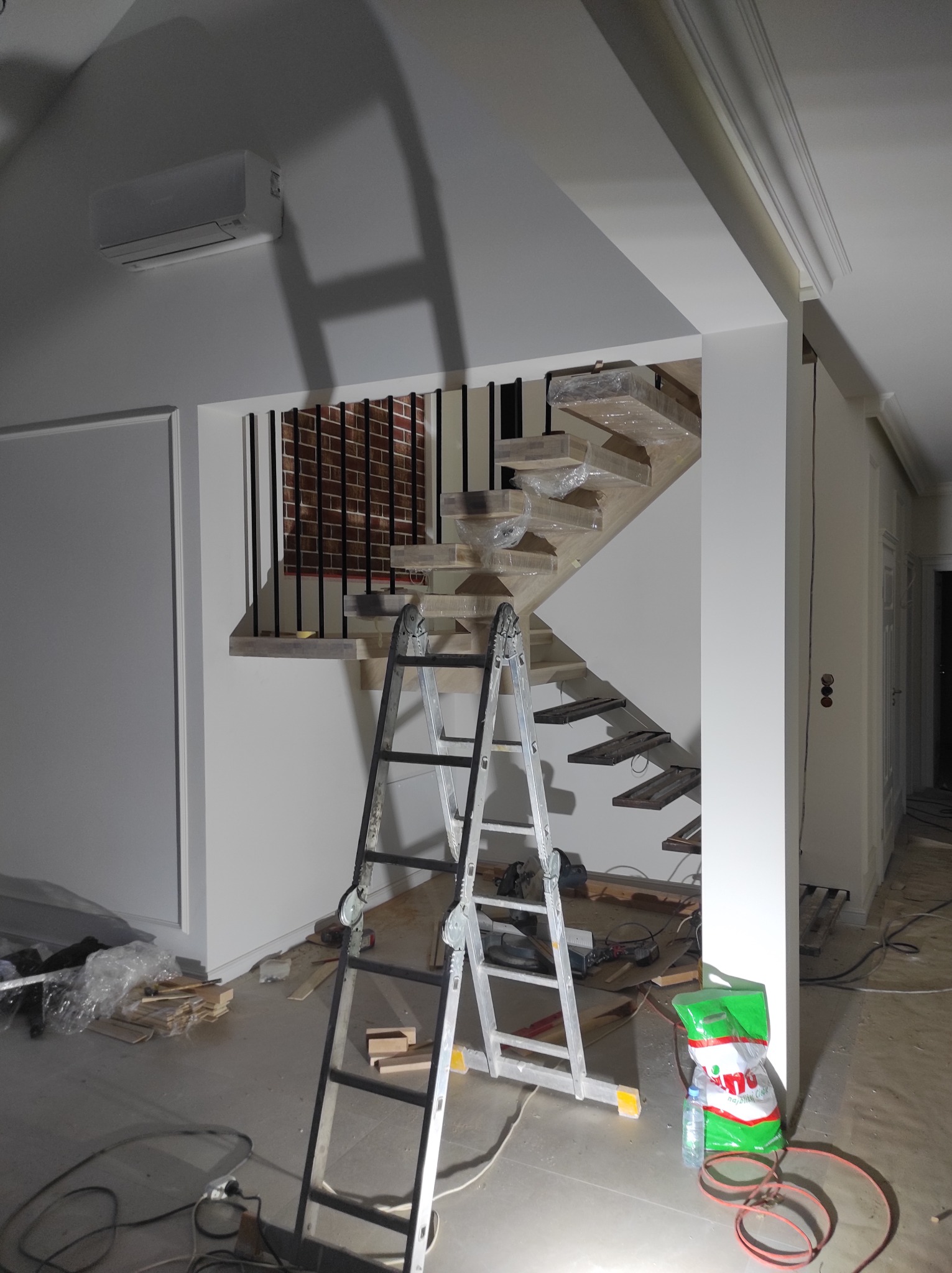 Wnętrze w trakcie wykańczania: drewniane schody tymczasowo zabezpieczone folią, aluminiowa drabina oparta o ścianę, widoczne materiały budowlane i narzędzia, ściany pomalowane na biało, zamontowany...