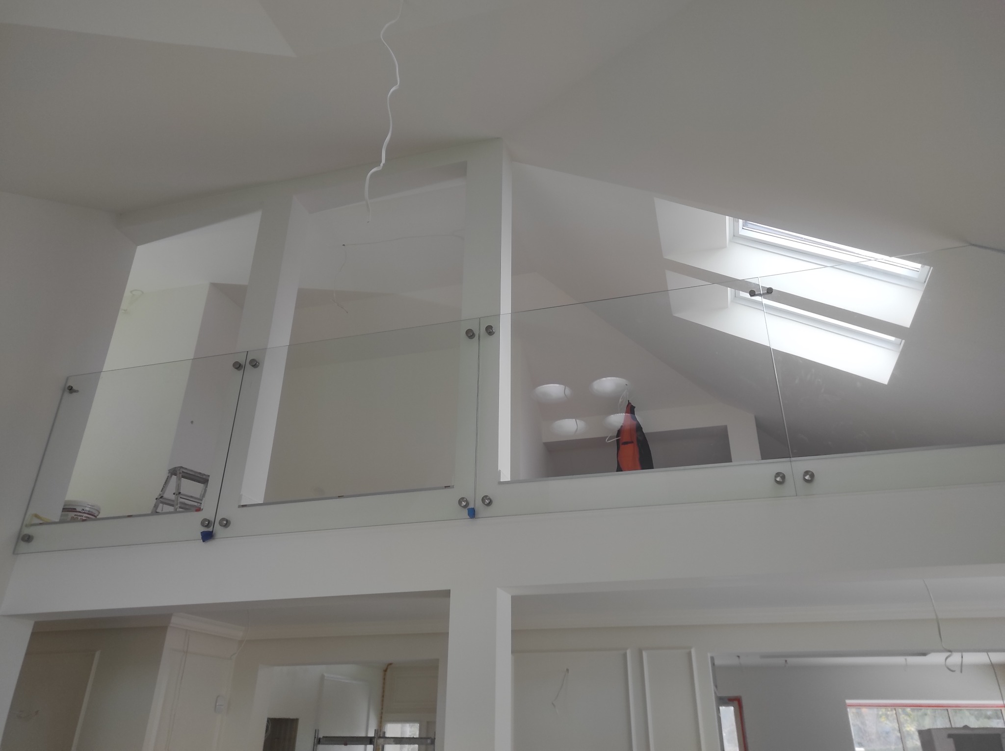 Nowoczesne, jasne wnętrze z antresolą zabezpieczoną szklaną balustradą mocowaną na metalowych uchwytach; widoczne okno dachowe i białe ściany.