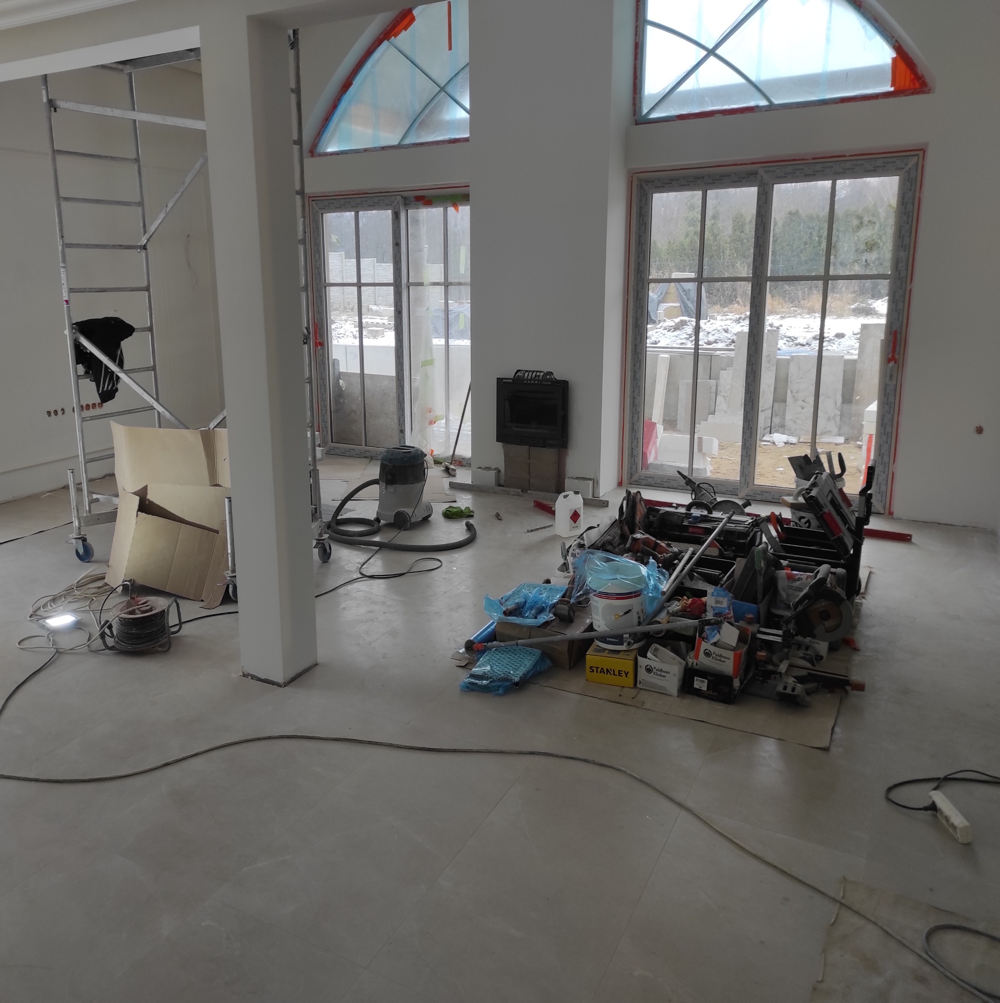 Wnętrze w trakcie prac wykończeniowych: widoczne szpachlowane ściany, świeżo położona podłoga z płytek, rusztowanie, okna zabezpieczone folią, kominek w trakcie obudowy oraz sterta narzędzi...
