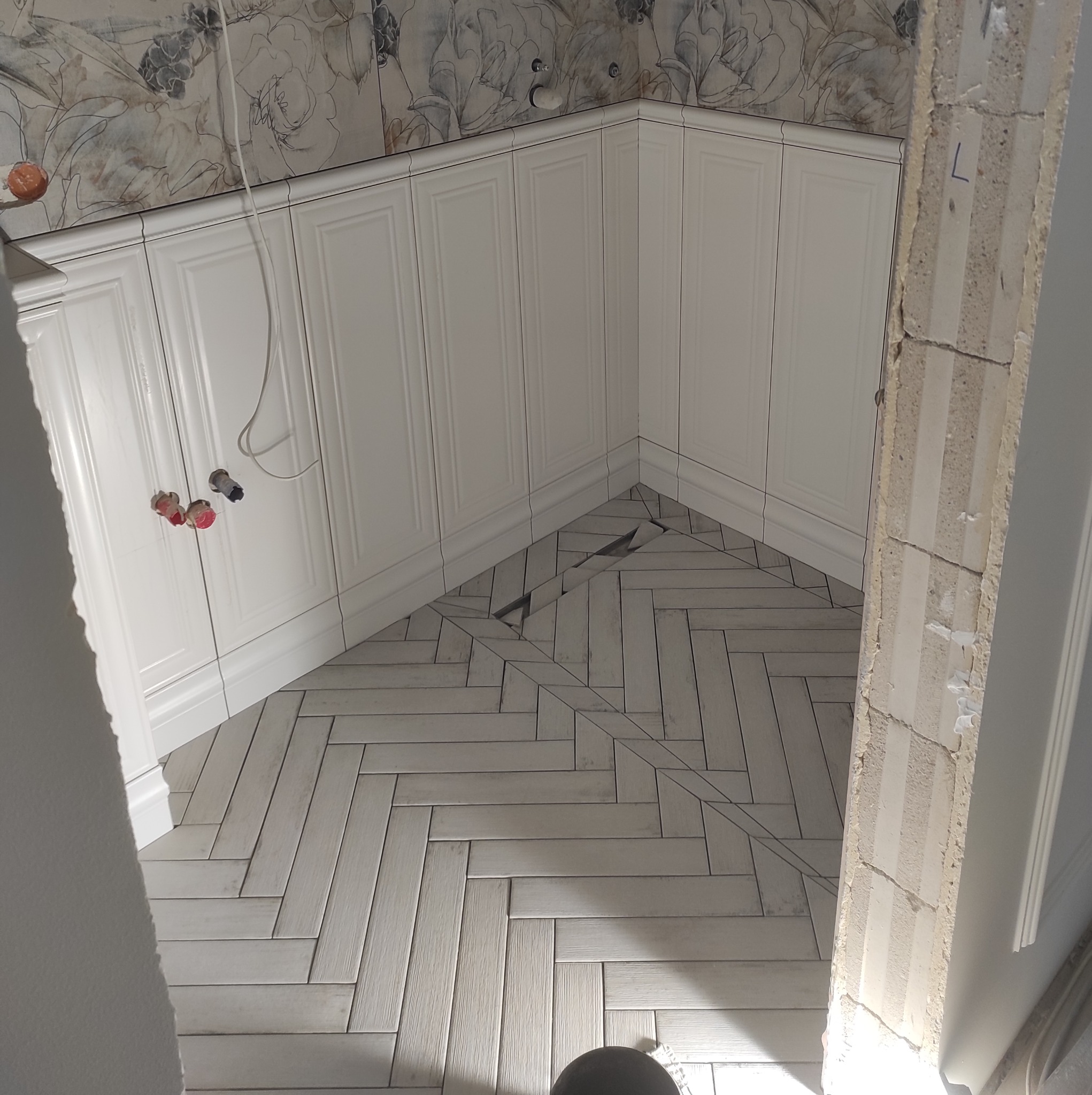 Białe szafki kuchenne z frezowanymi frontami i ułożona w jodełkę podłoga z płytek imitujących drewno, widoczne fragmenty instalacji wodnej i fragment ściany z dekoracyjną tapetą w kwiaty podczas...