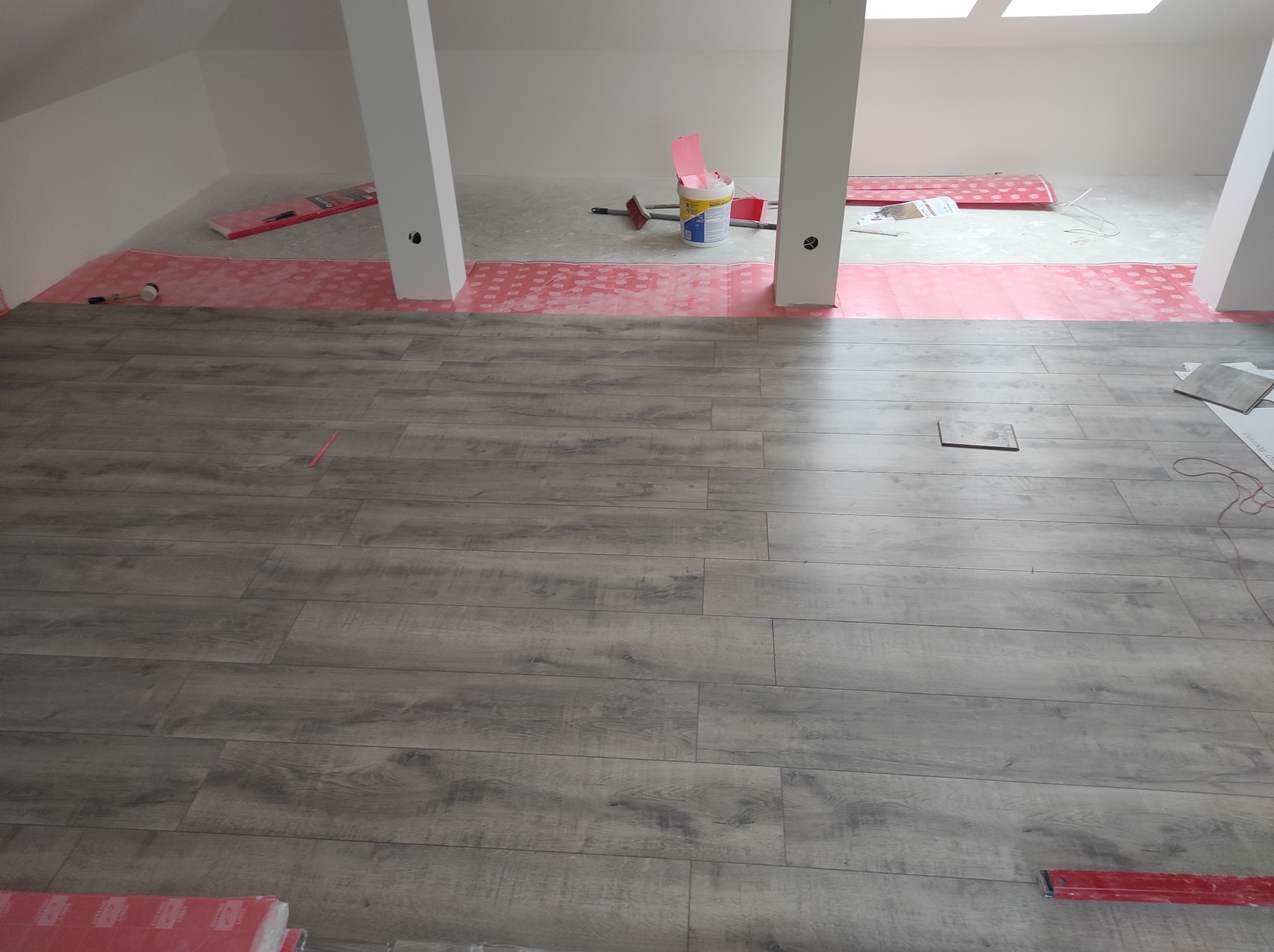 Układanie paneli podłogowych w szarym odcieniu, widoczne narzędzia, podkład i puszka z farbą w surowym wnętrzu poddasza z kolumnami.