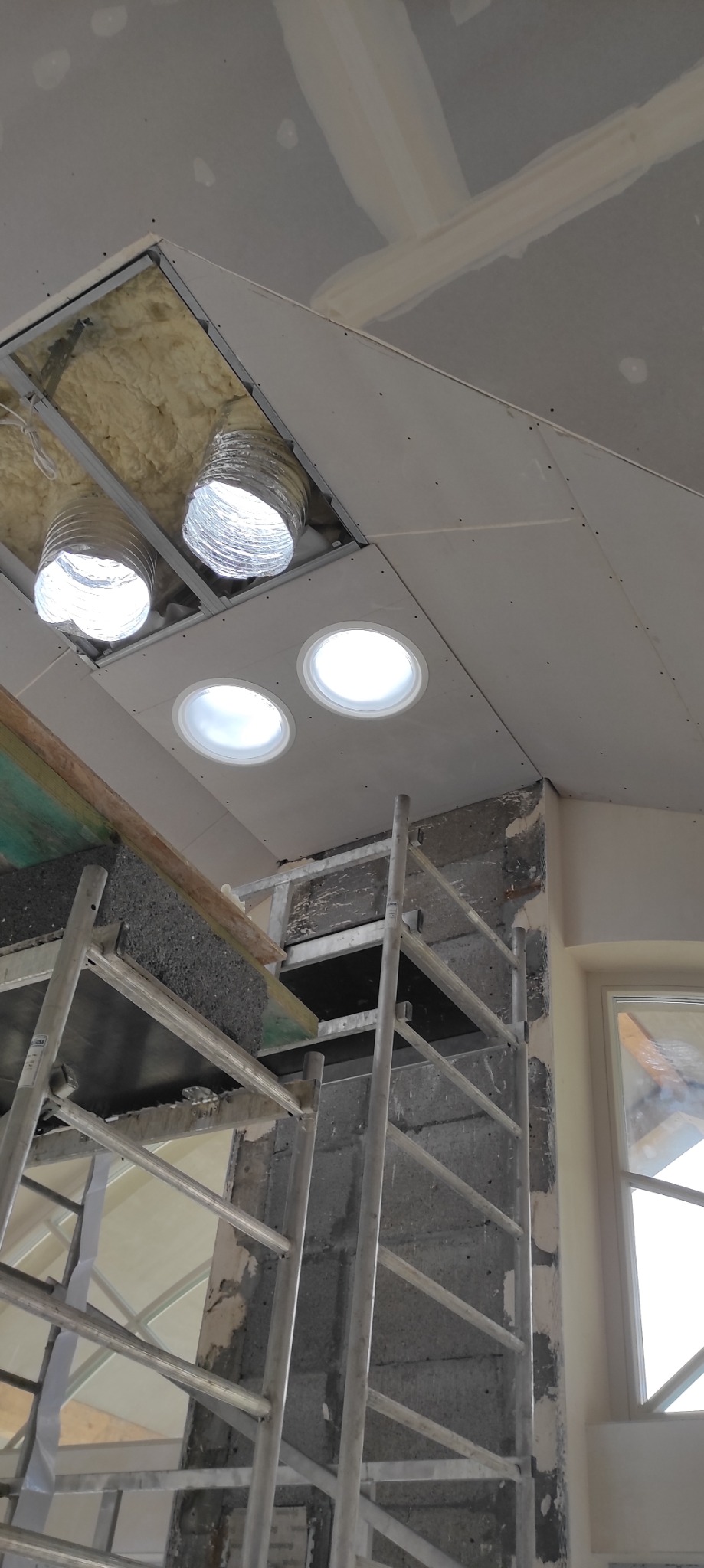 Montaż sufitu podwieszanego z oknami dachowymi typu świetlik tunelowy, widoczne elementy konstrukcyjne i izolacja, rusztowanie w pomieszczeniu.