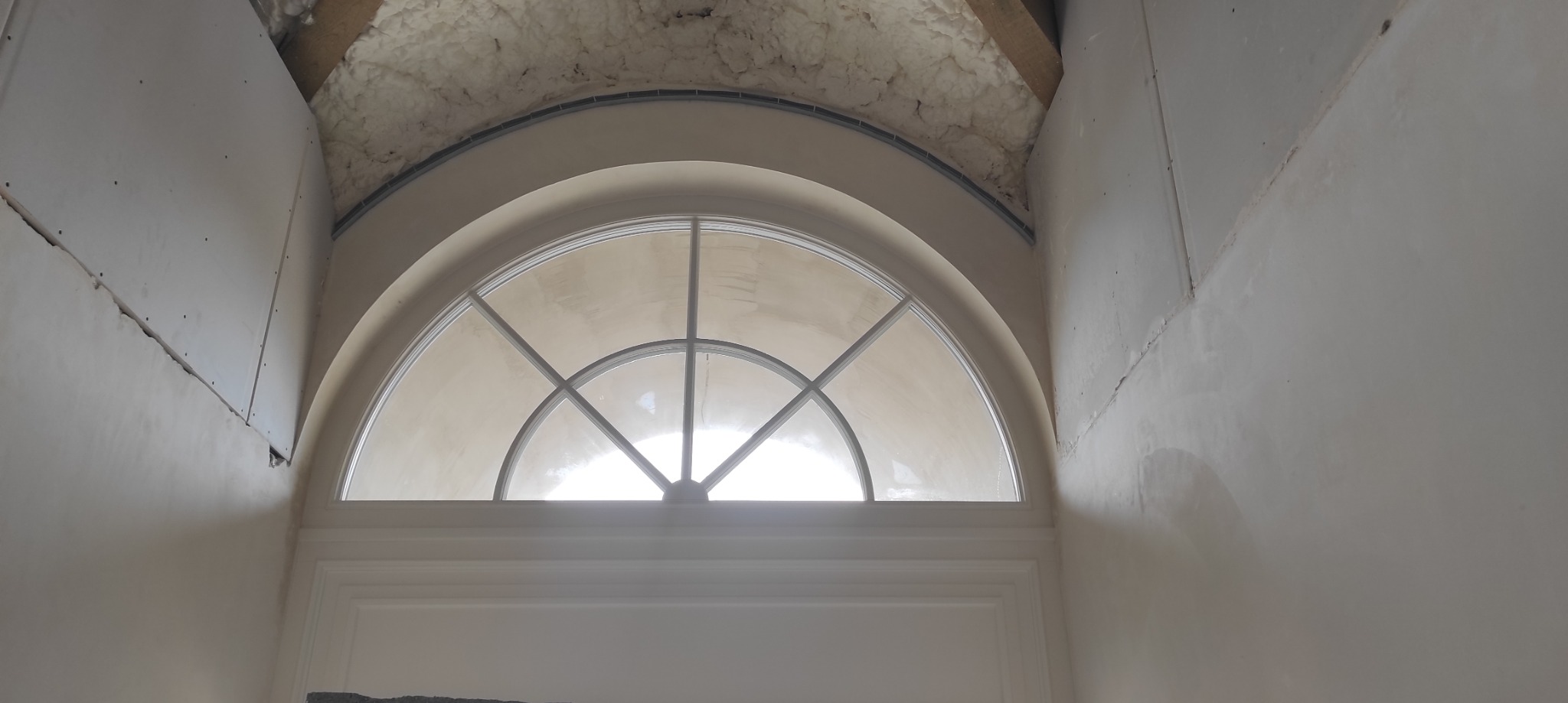 Półokrągłe okno z białymi szprosami w trakcie montażu, widoczne płyty gipsowo-kartonowe i izolacja stropu, ściany w trakcie szpachlowania.